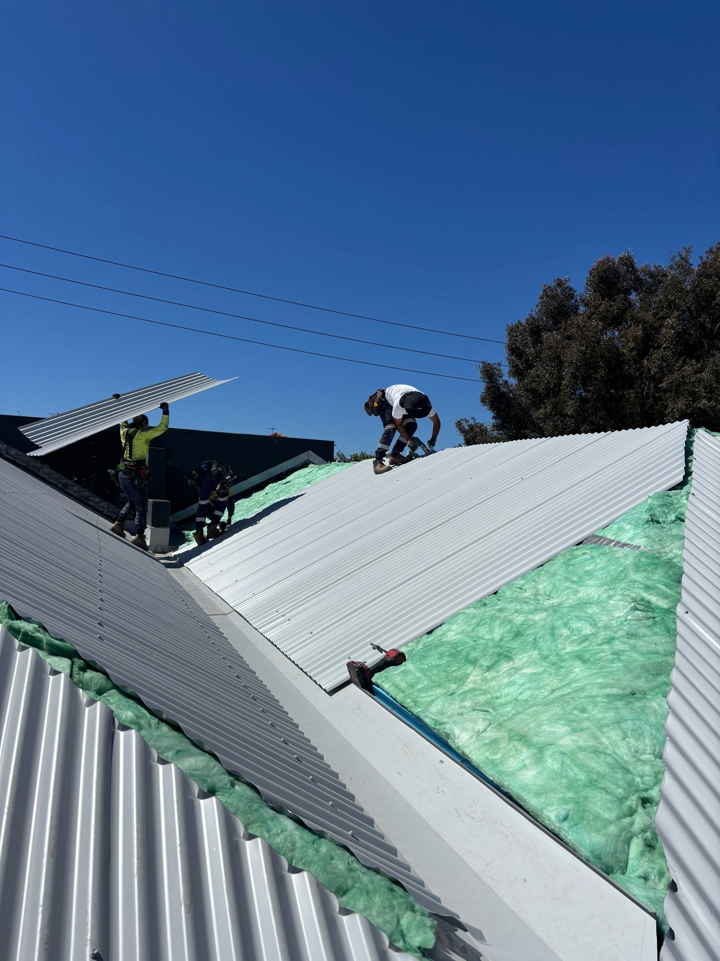 Construction workers installing metal roofing on a building, with insulation visible underneath. One worker is kneeling on the roof, while others are handling roofing sheets, under a clear blue sky.