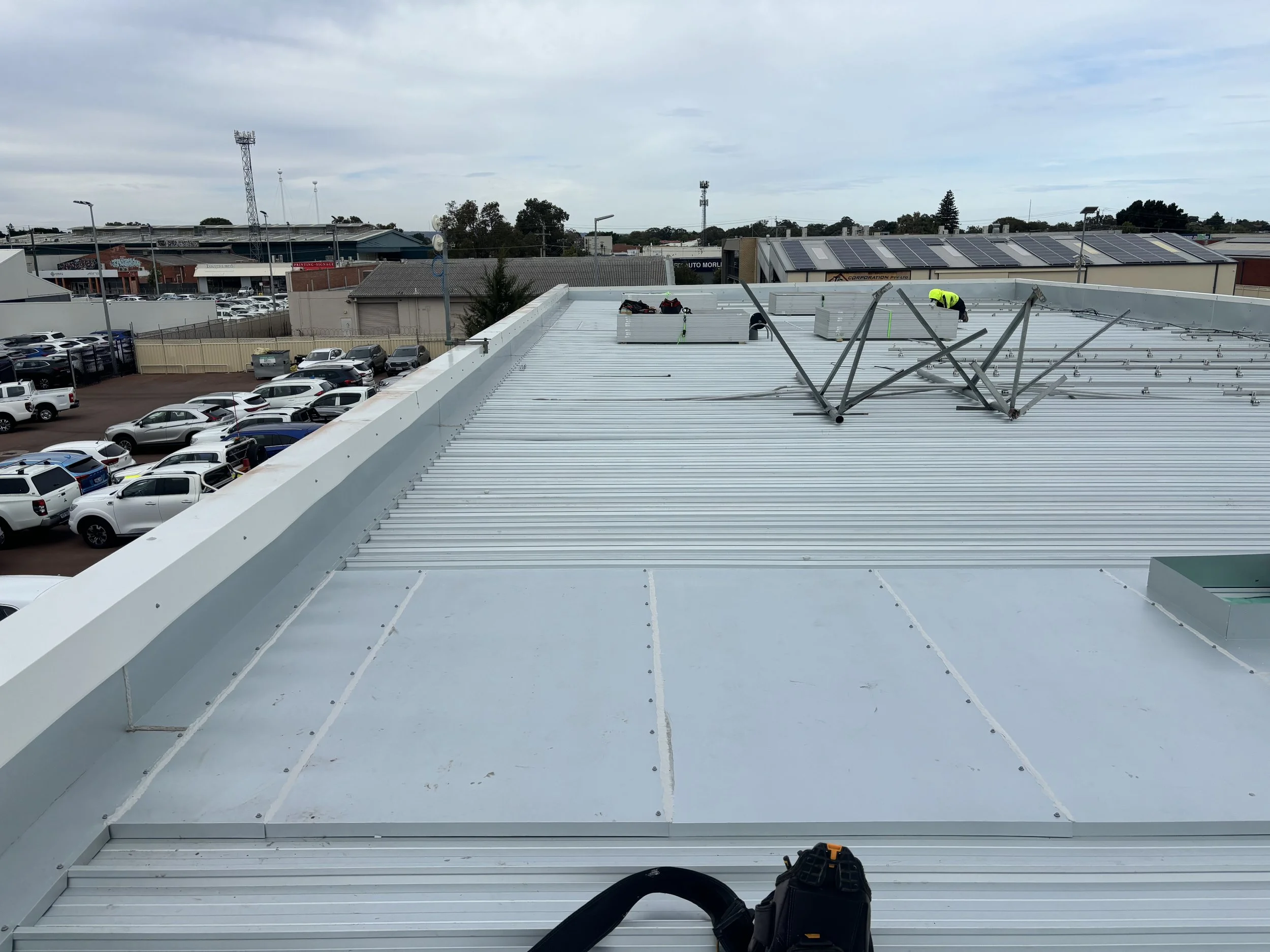 Workers installing or repairing a metal roof on a commercial building with a parking lot full of cars in the background.