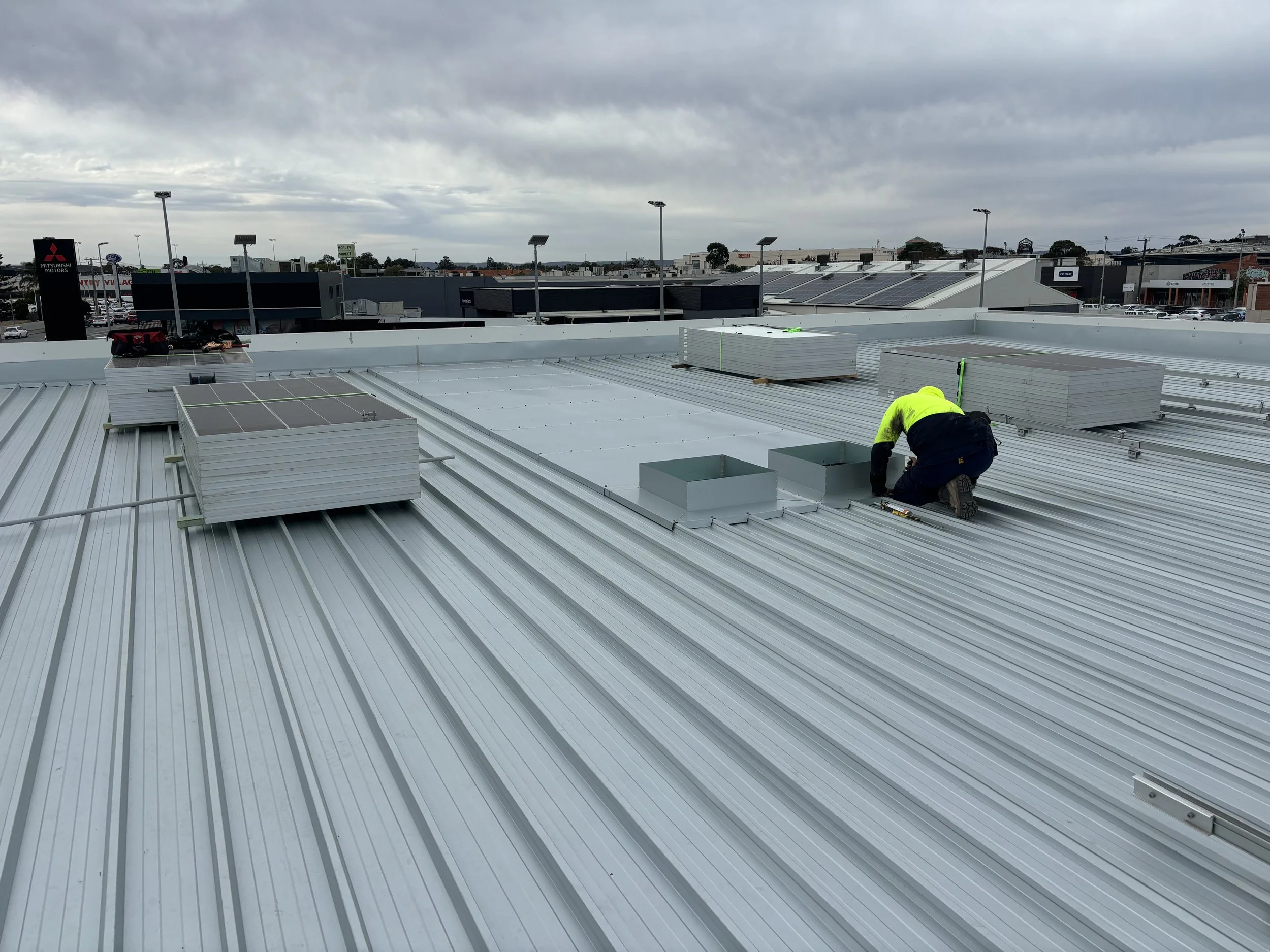 A construction worker in a yellow safety vest kneels on a metal roof, working on vents and panels during an overcast day.