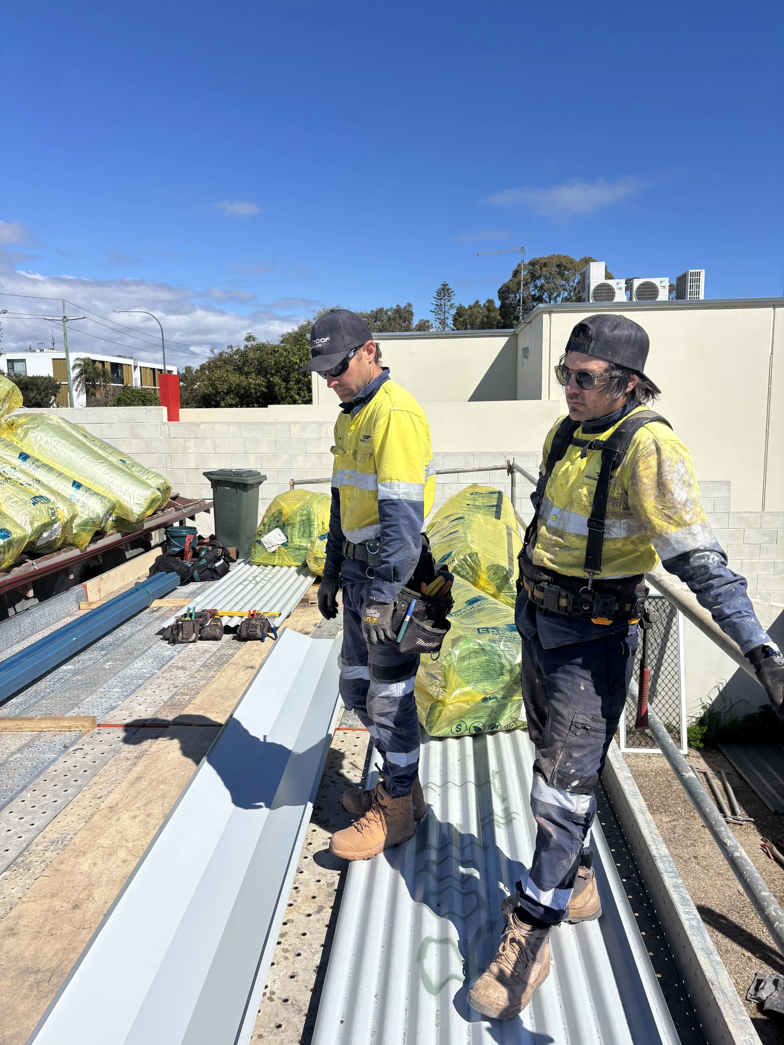 Two construction workers in high-visibility jackets and work boots standing on a metal roof at a construction site, surrounded by construction materials and tools, with a clear blue sky overhead.