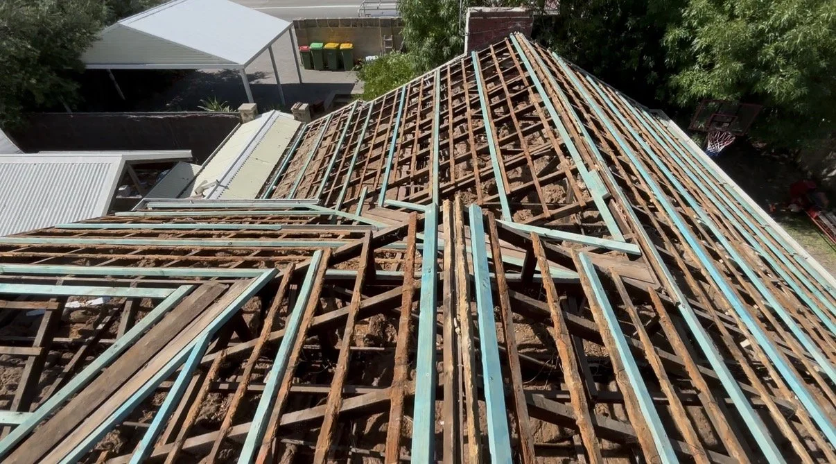 View from the top of a building under construction shows exposed wooden and metal framing for the roof, with a partial roof structure on the left side and surrounding trees and structures nearby.