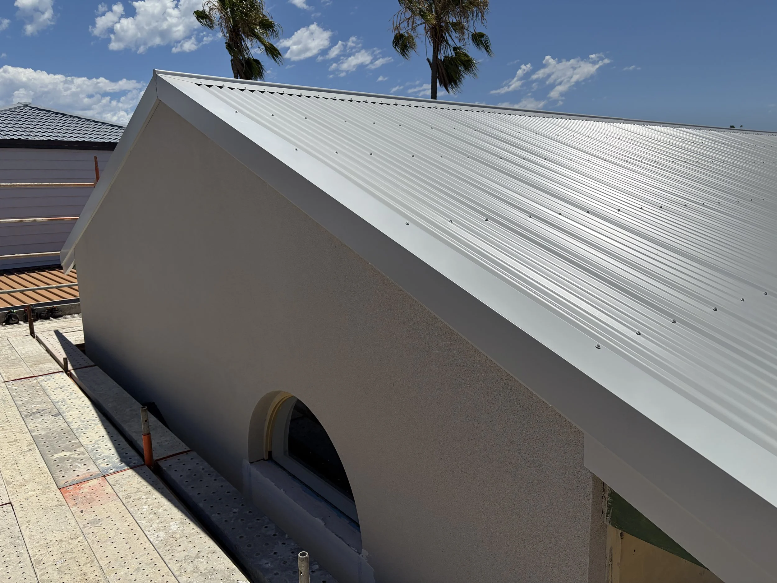 A close-up view of a house roof under construction with a metal roof panel and part of a flat wall with a small arched window, with blue sky and palm trees in the background.