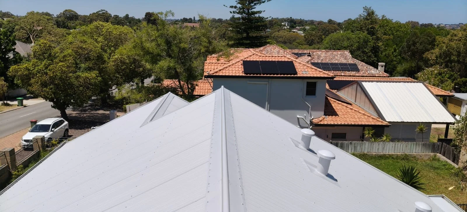 View of rooftops with solar panels, trees, and a street with a parked car in a suburban neighborhood.