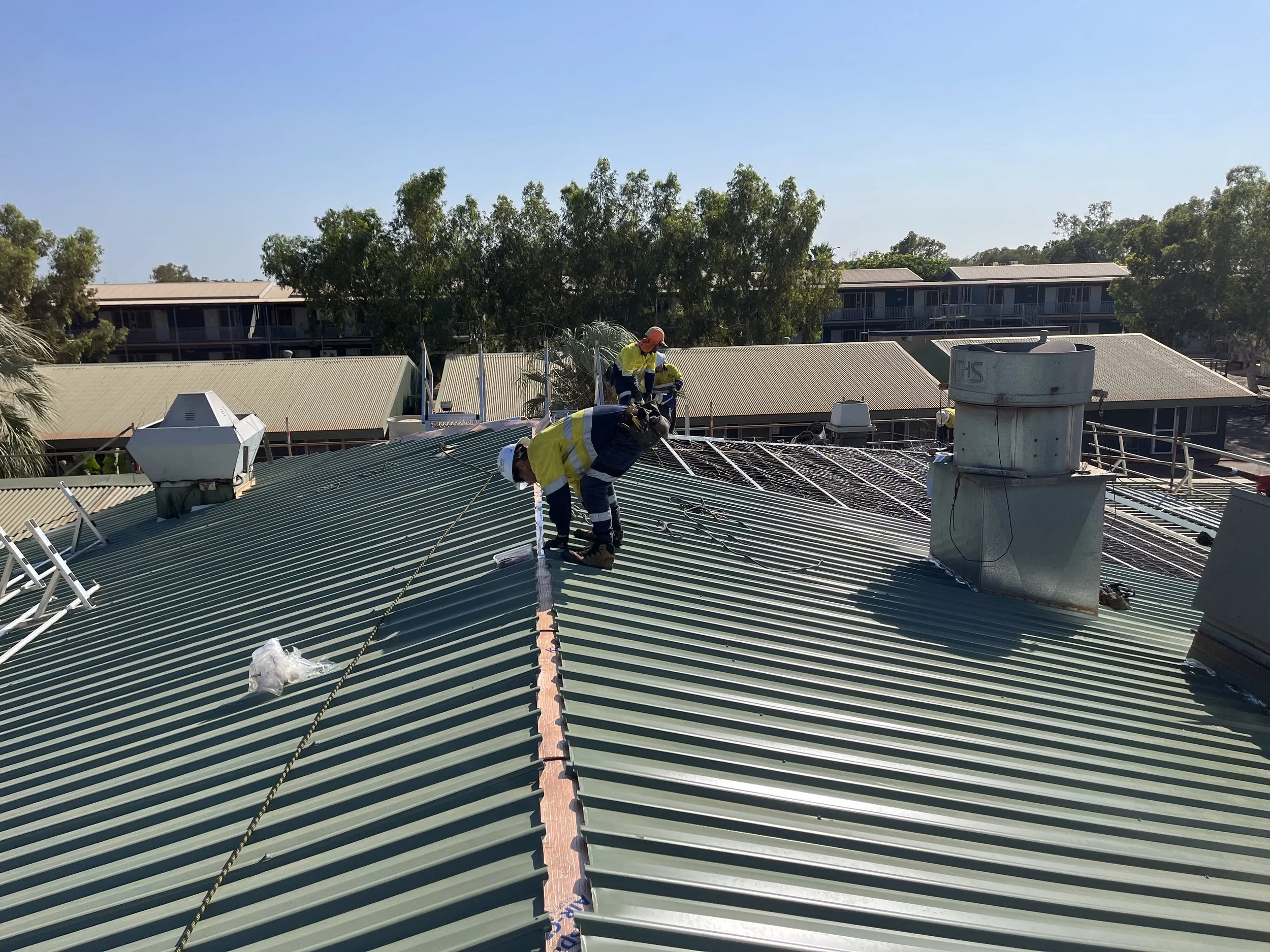 Construction workers in safety vests and helmets working on a metal roof on a sunny day.