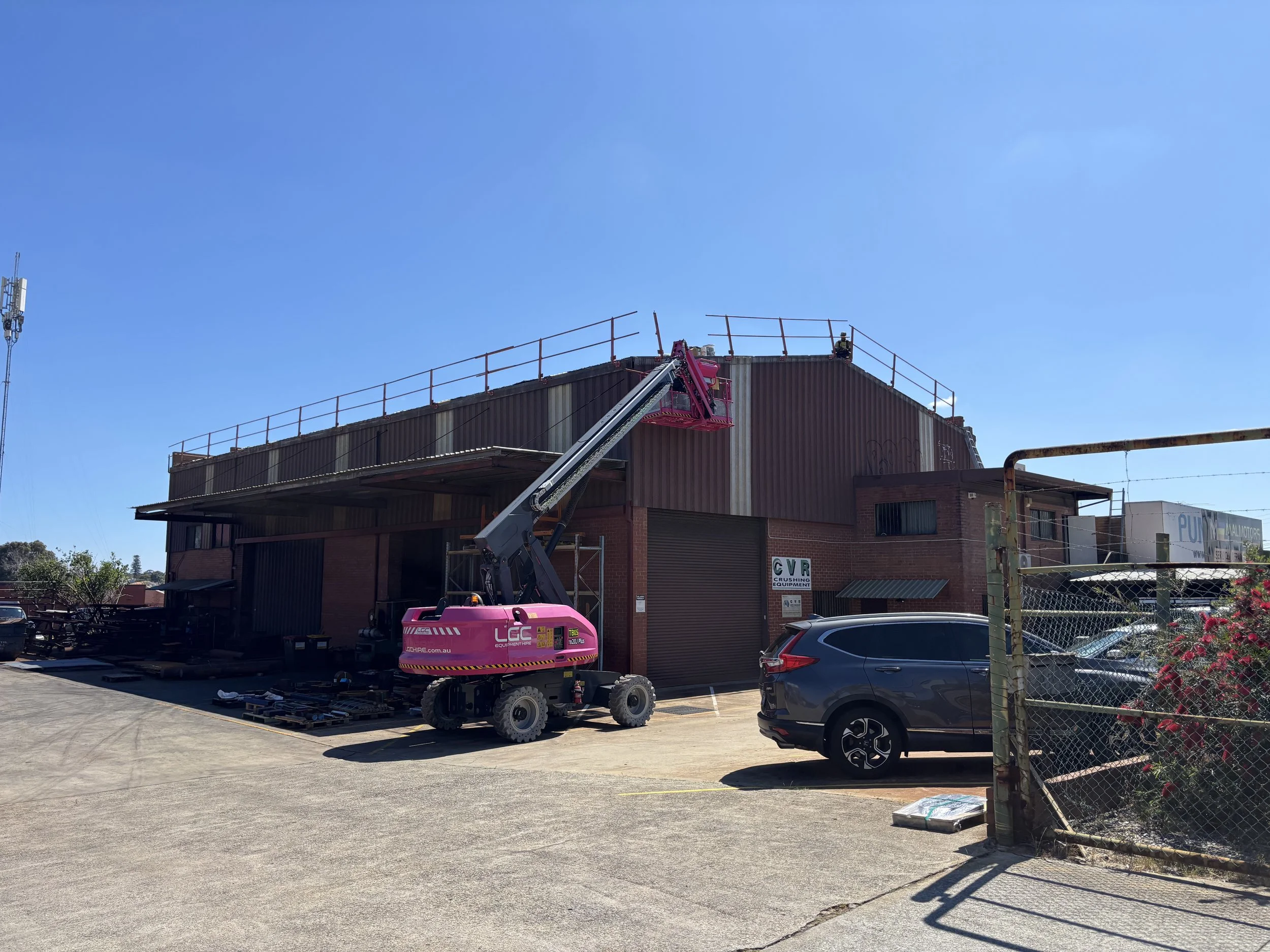 A pink telescopic boom lift extending towards the roof of a brick commercial building under construction or repairs, with a gray and black car parked nearby and a fenced area with pink flowers in the foreground.