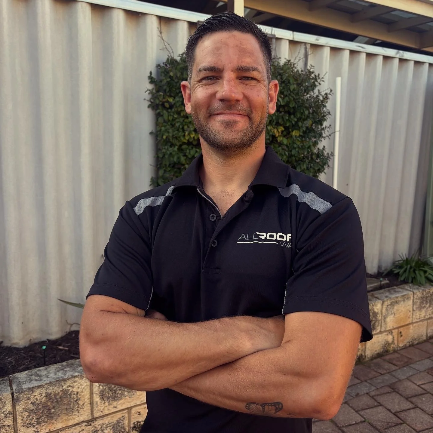 A man with dark hair and a beard standing outside with arms crossed, wearing a black polo shirt with the logo 'ALLROOF' in white, in front of a white fence and greenery.