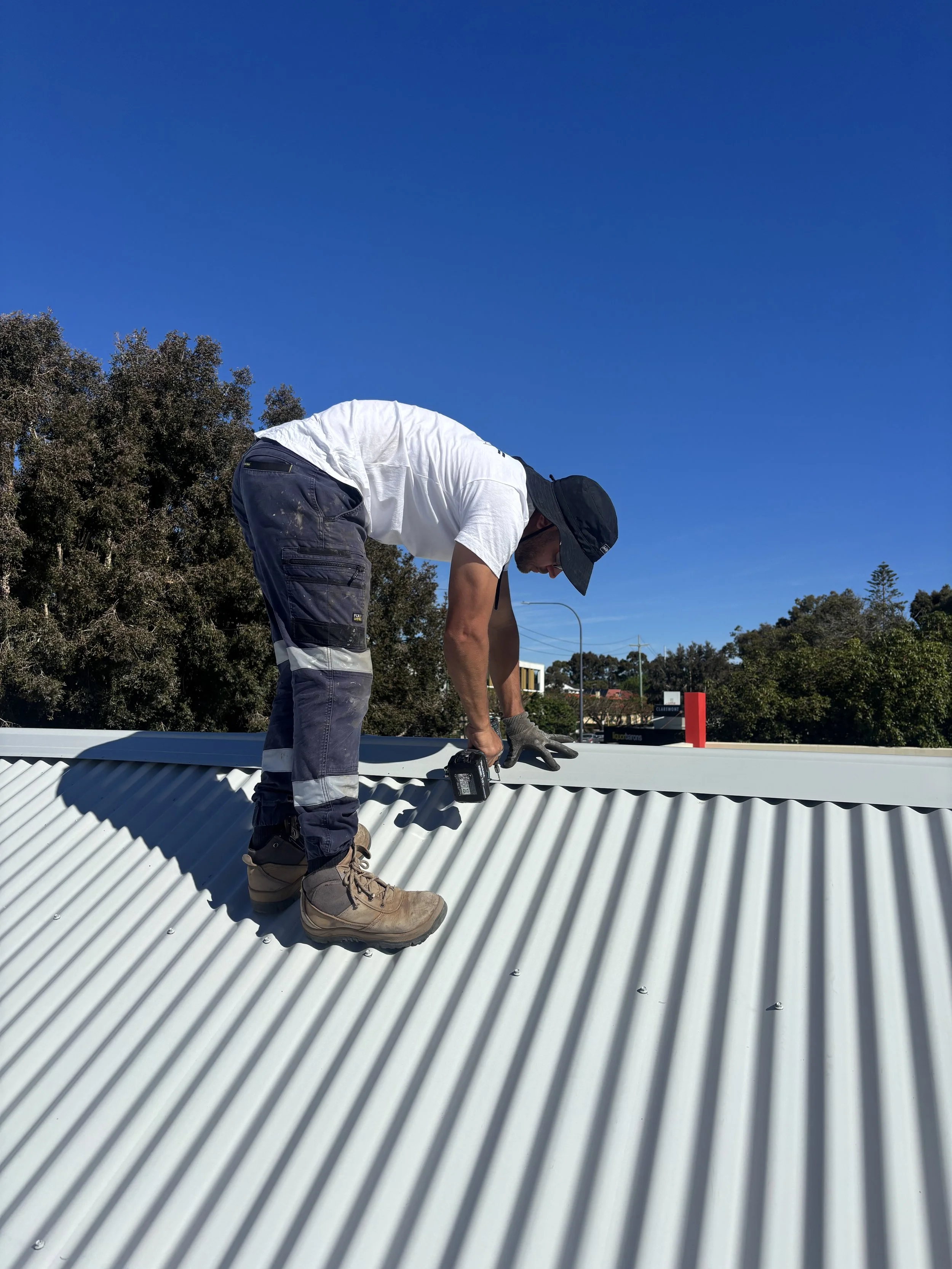 Worker in white shirt, dark pants, tan boots, black hat, and gloves installing or inspecting a corrugated metal roof on a sunny day, with trees and blue sky in the background.