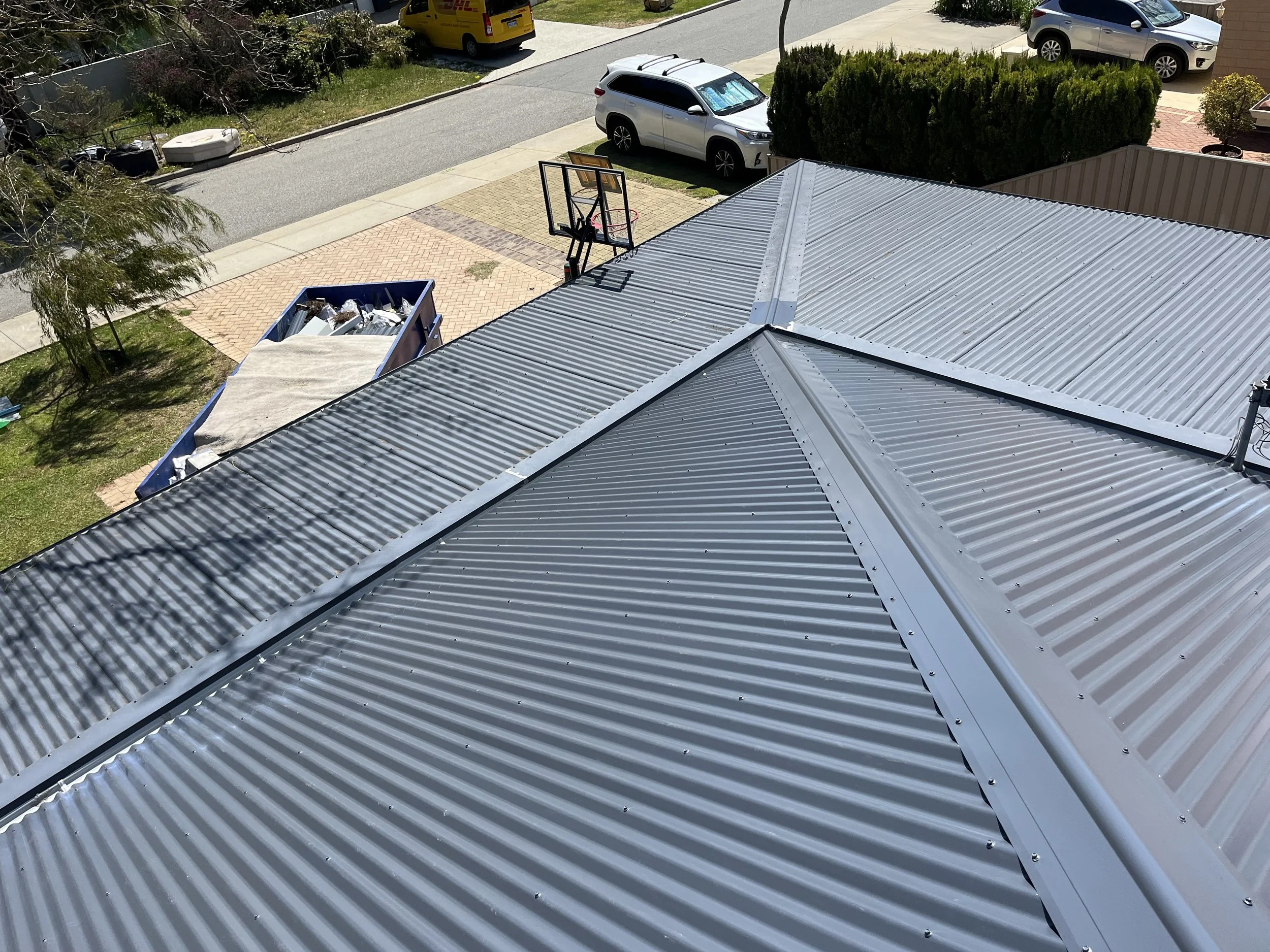 View from above of a metal roof with ridge vents and a basketball hoop mounted on the roof structure. Cars parked on the street and a trash bin with debris are visible in the background.