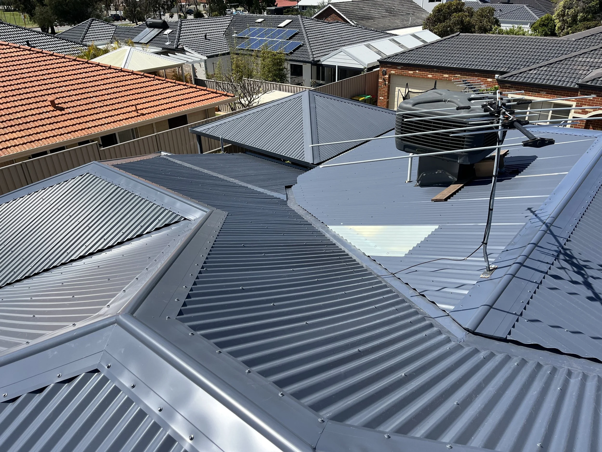 View of metal roofs on multiple houses in a neighborhood, with a rooftop air conditioning unit and TV antenna visible in the foreground.