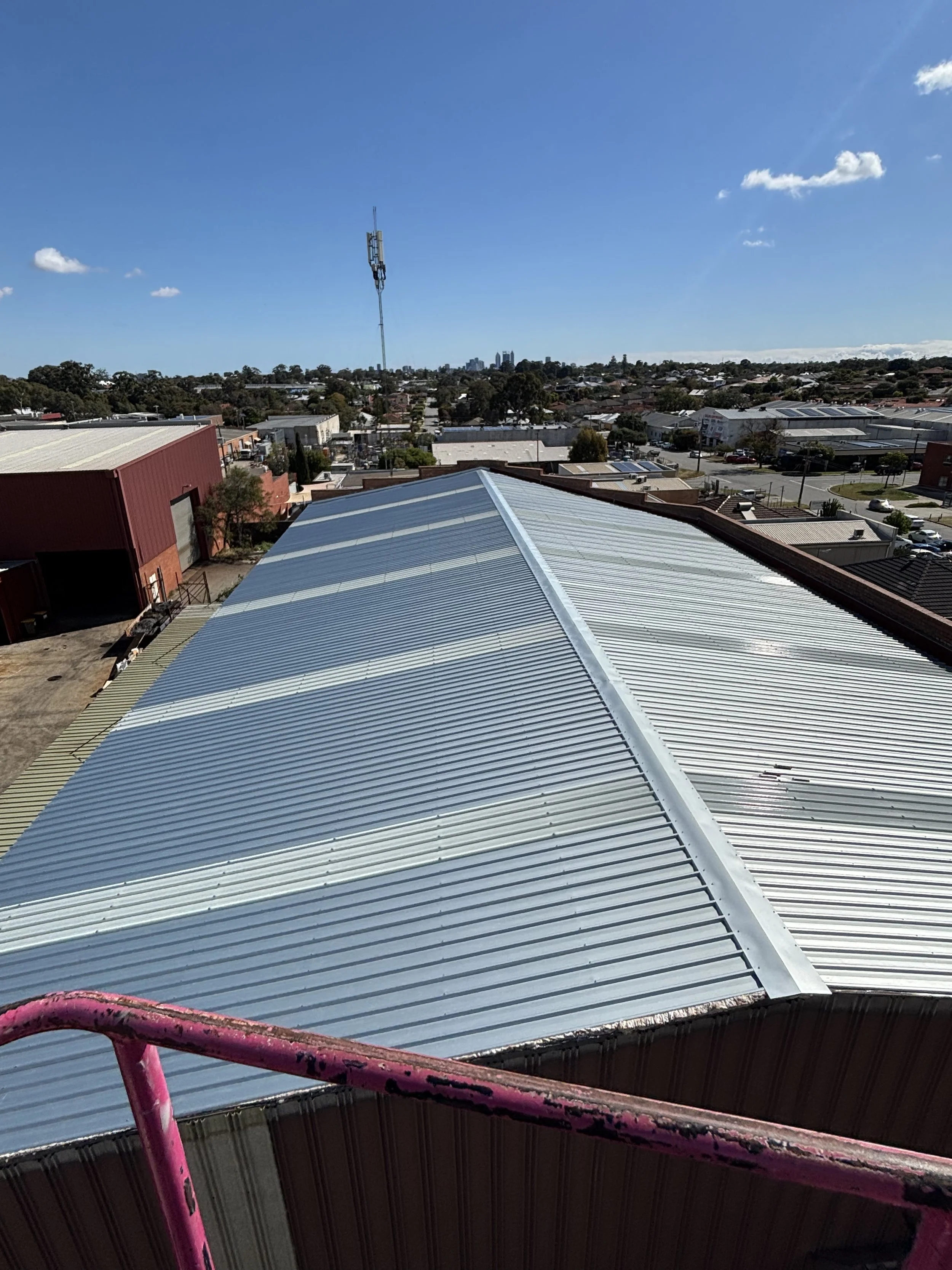A metallic corrugated roof on a commercial building under a clear blue sky with a few clouds, viewed from a high vantage point.