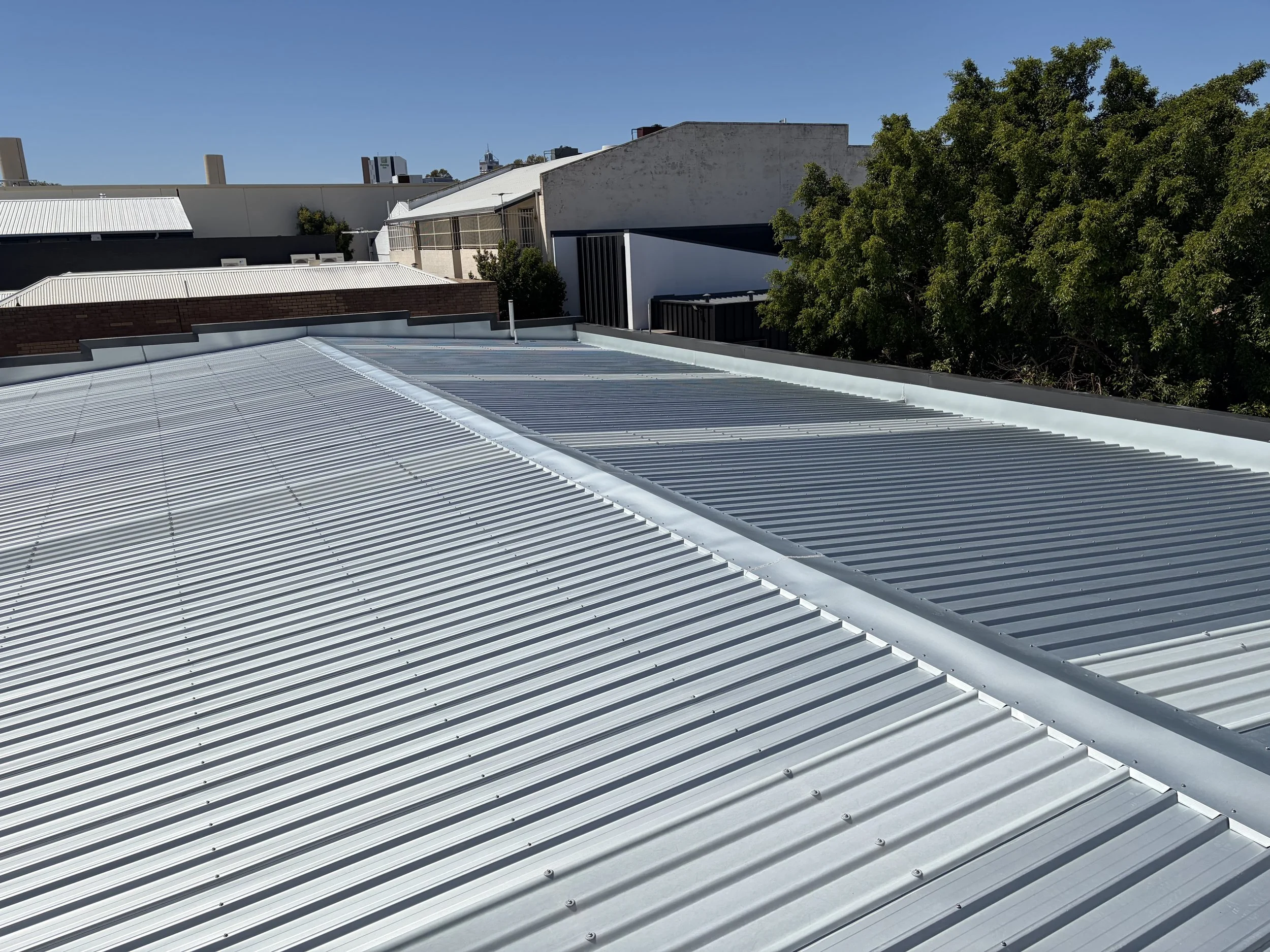 View of a metal rooftop with ridges, some sections are shiny and new, in an urban area with buildings and trees under a clear blue sky.
