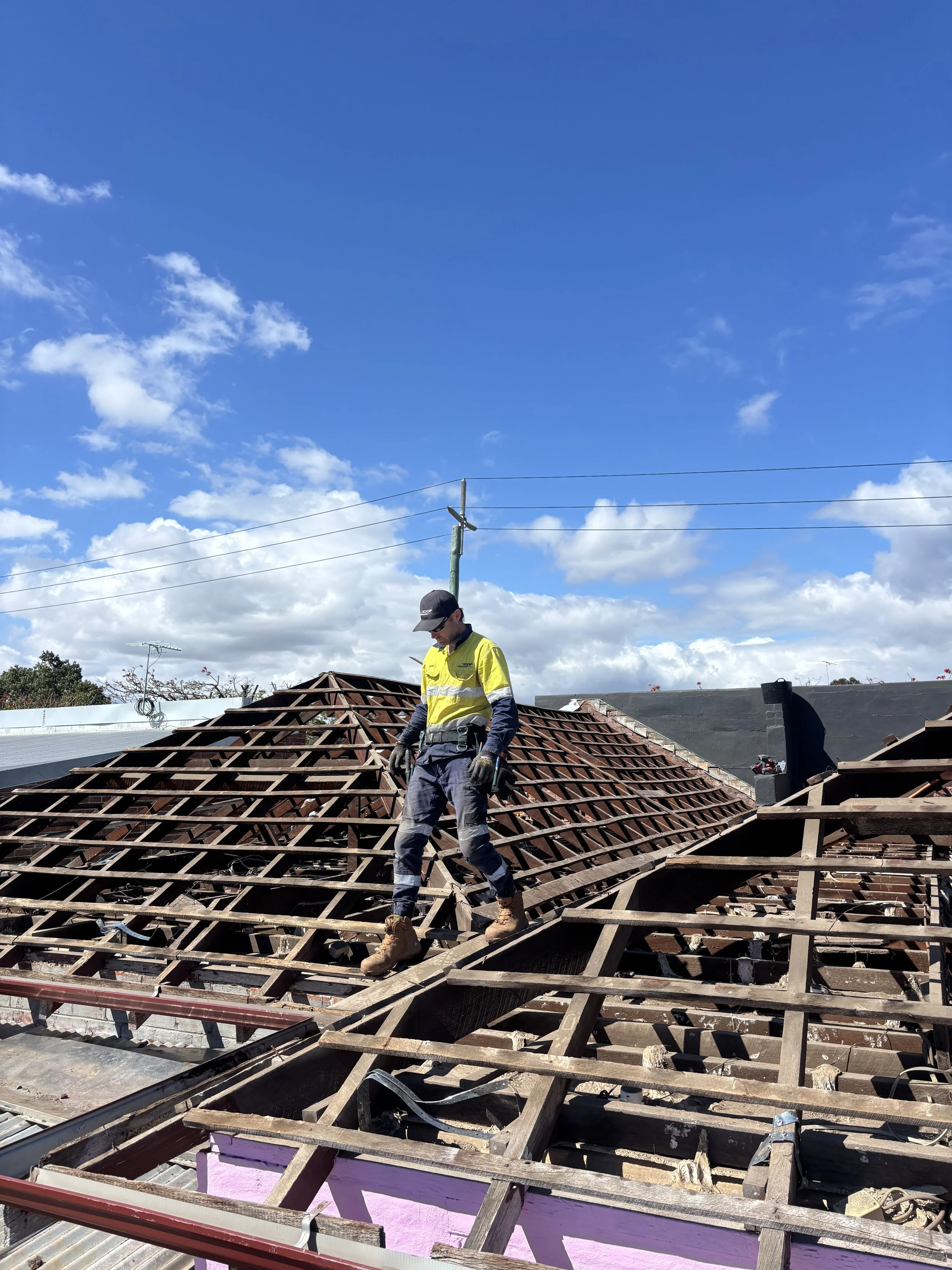 A worker in safety gear walking on a damaged roof with exposed wooden beams, under a blue sky with clouds.
