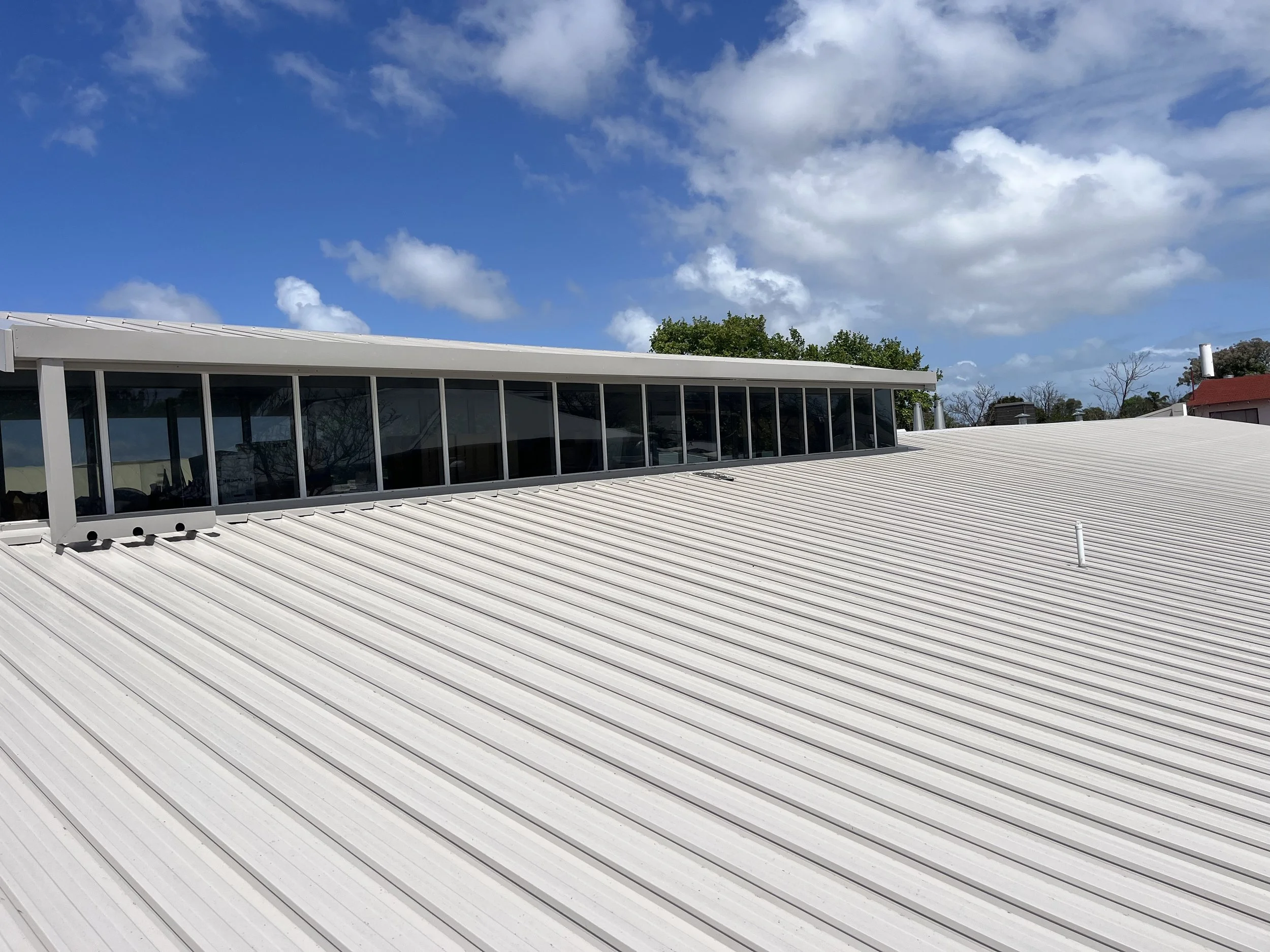 The image shows a white metal roof with a large rectangular glass section near the top, under a partly cloudy blue sky, with some trees and buildings visible in the background.