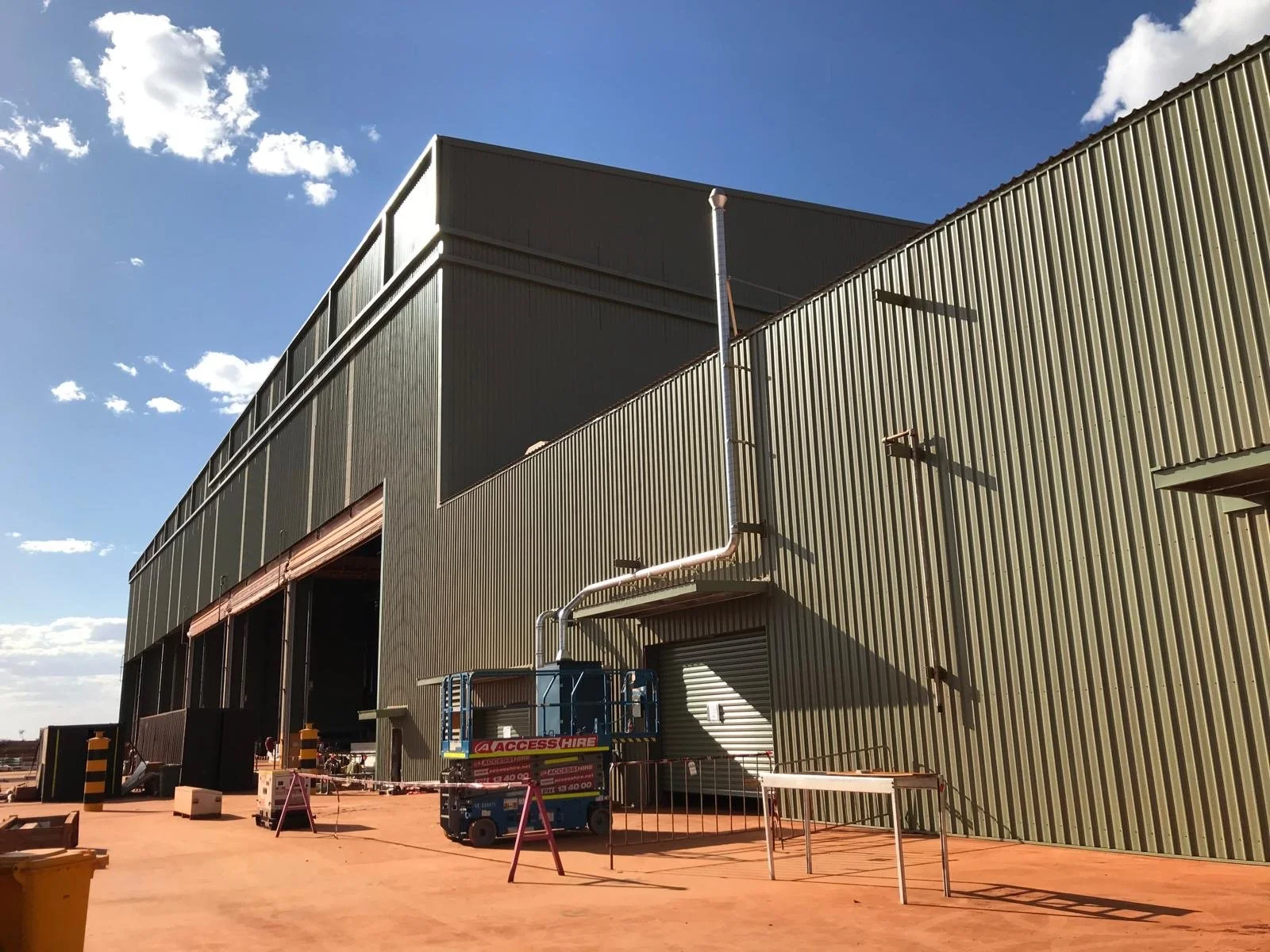 Exterior view of a large industrial building with corrugated metal walls, a lifted section, and construction equipment nearby under a partly cloudy sky.