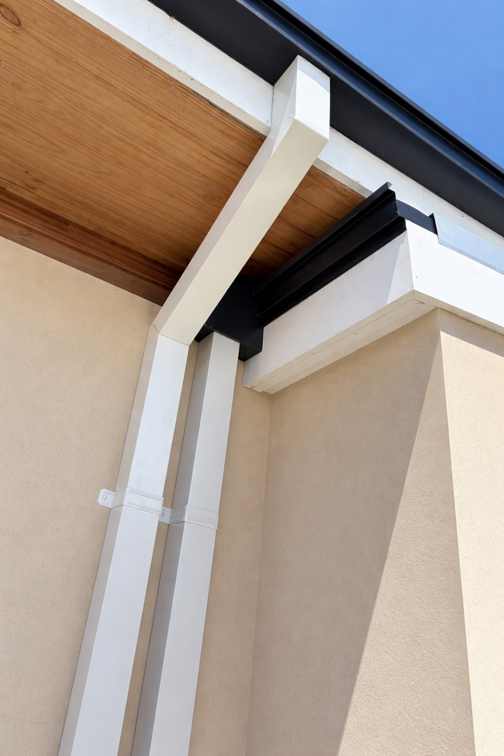 Close-up of the corner of a modern house exterior, showing beige stucco wall, white metal pipes, and a wooden ceiling soffit with black gutters and a clear blue sky.