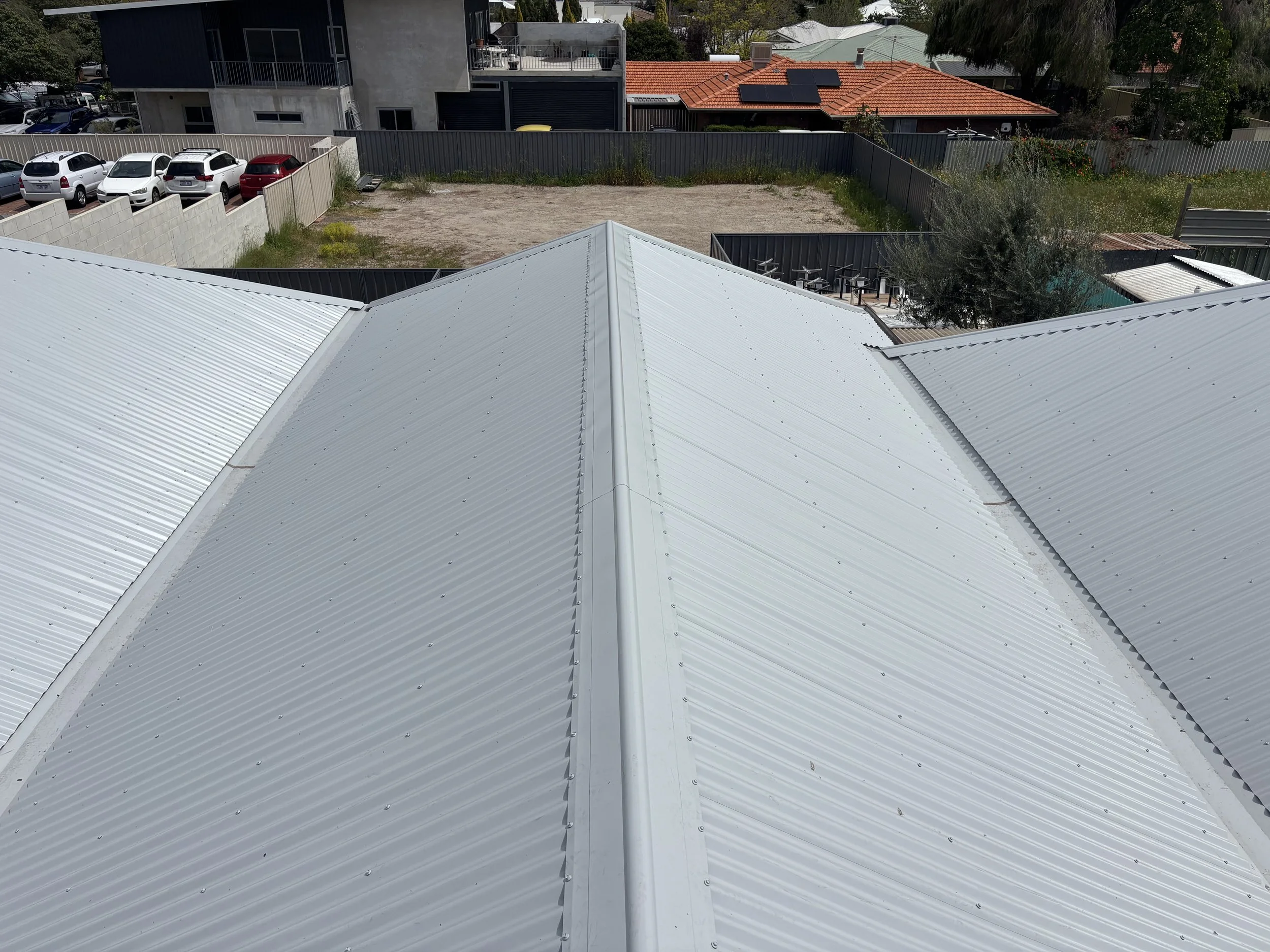 View of a metal roof with parallel ridges, seen from above, with a yard and houses in the background.