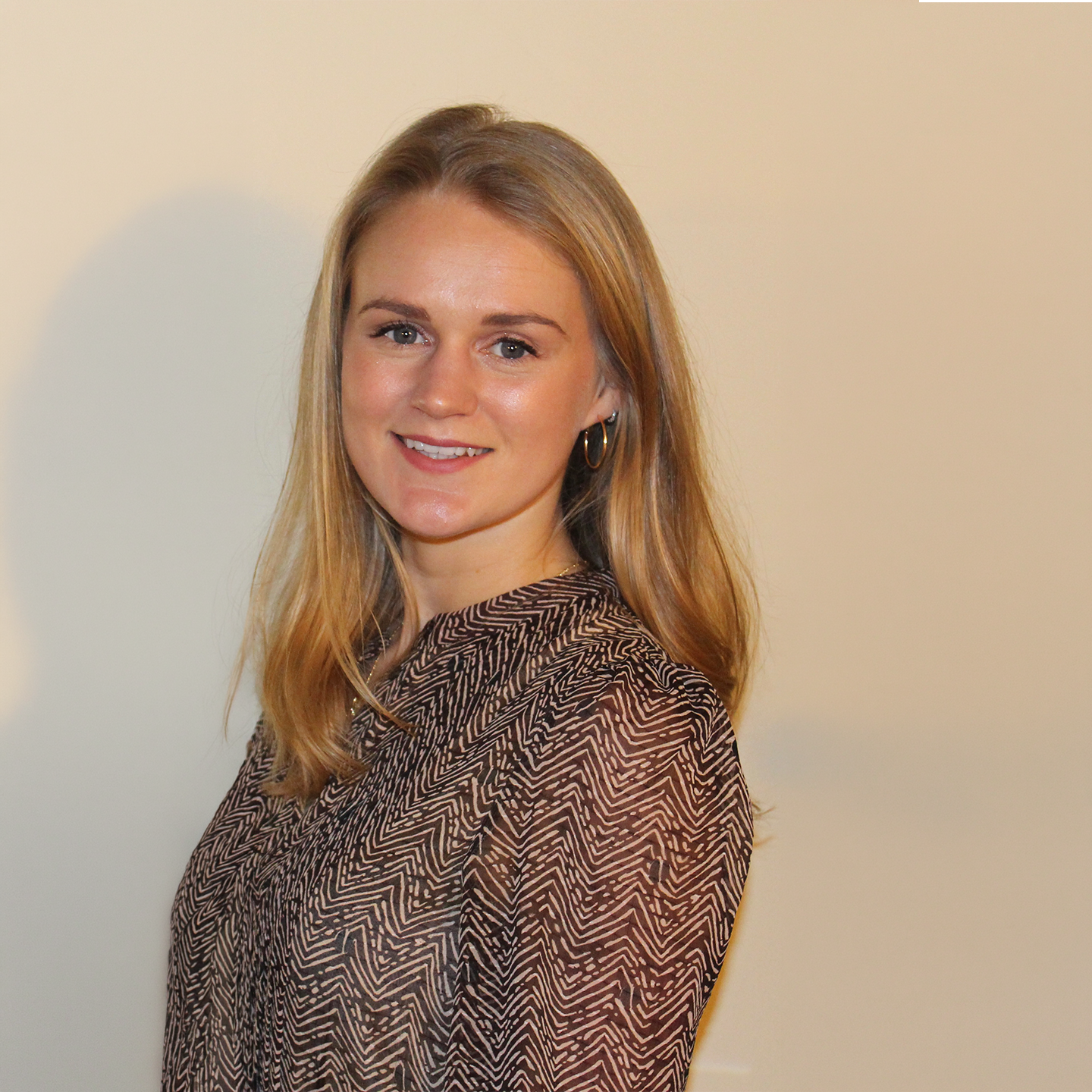 A young woman with blonde hair, smiling, wearing a patterned blouse and hoop earrings, standing against a plain cream-colored background.