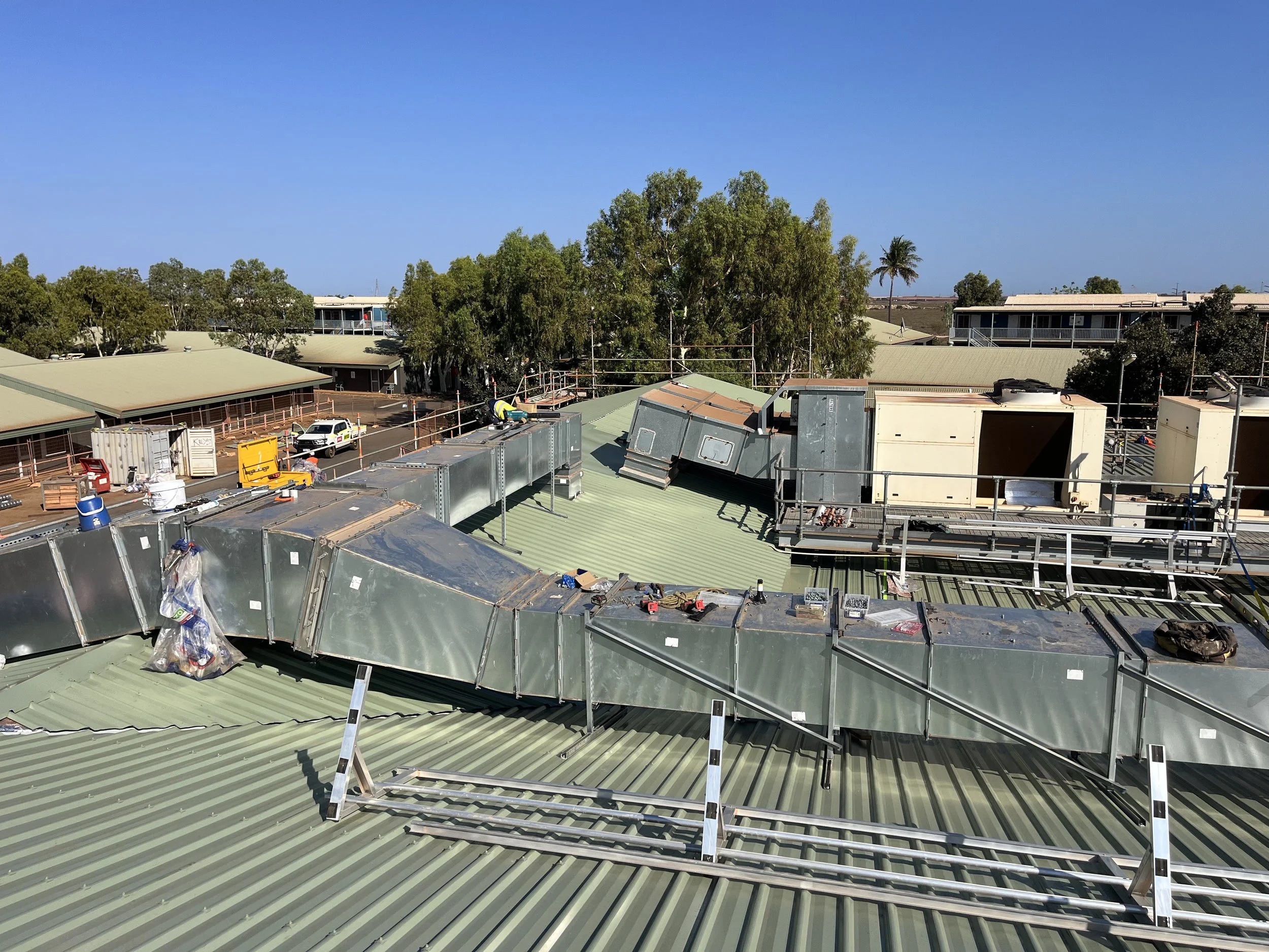 Construction workers installing ductwork on a green metal roof in a commercial building, with trees and other buildings in the background.