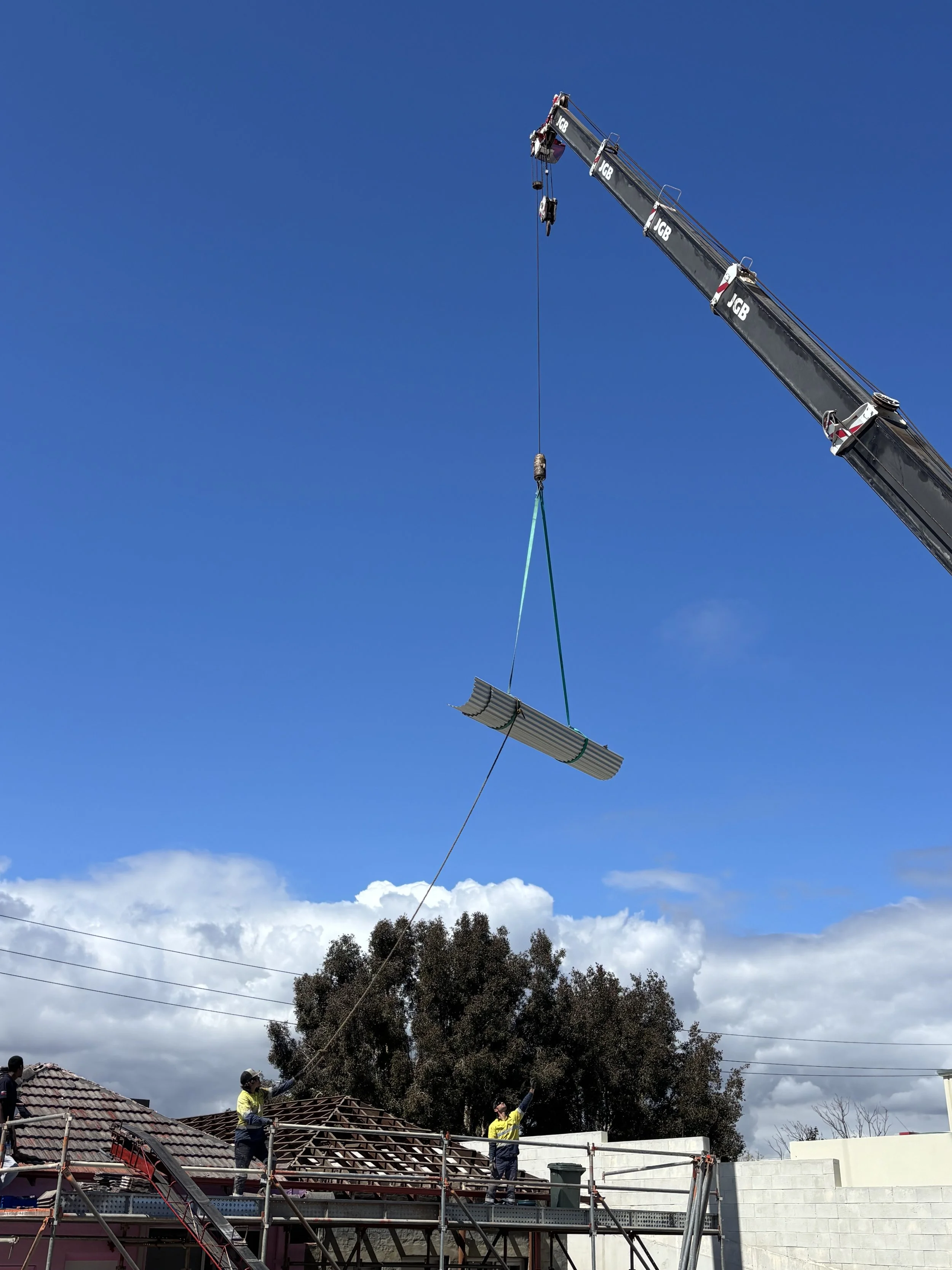 Construction workers raise a metal roof panel with a crane on a building site during daytime.