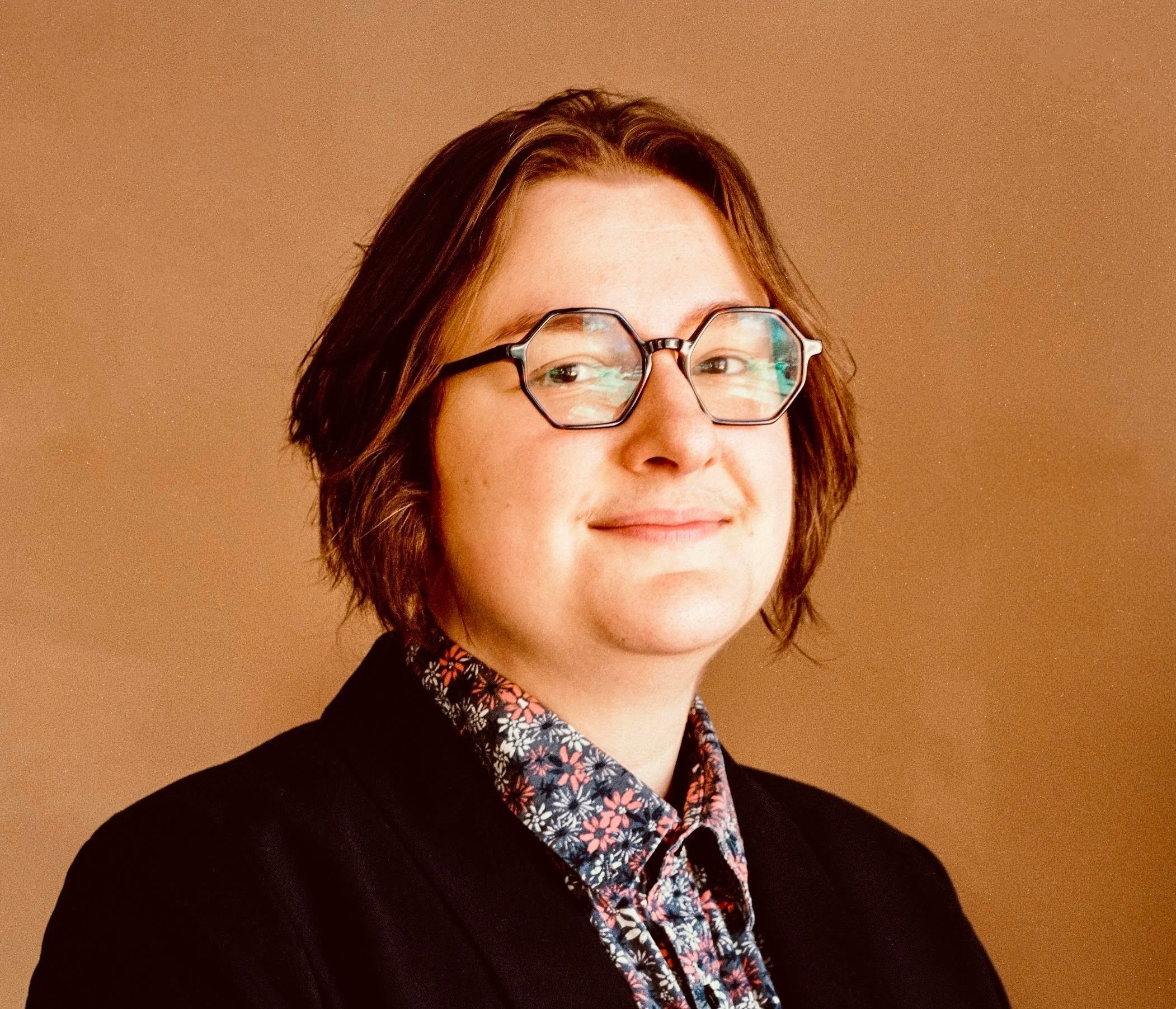 A photo of Ash, a person wearing dark rimmed glasses, pink, a white, and blue button-up, with medium length hair and a black blazer