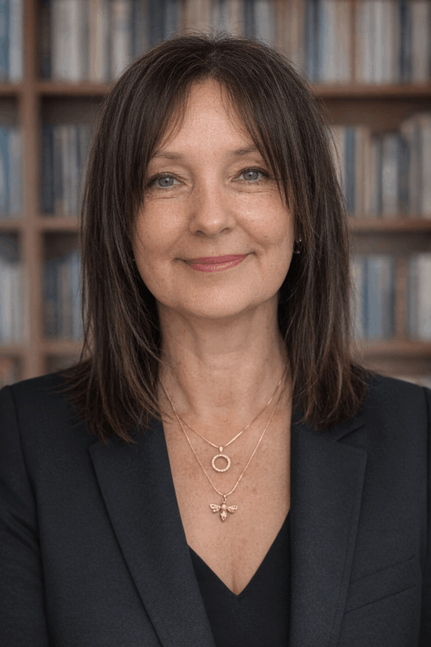 A woman with shoulder-length brown hair and blue eyes wearing a black blazer and layered necklaces, standing in front of a bookshelf filled with books.