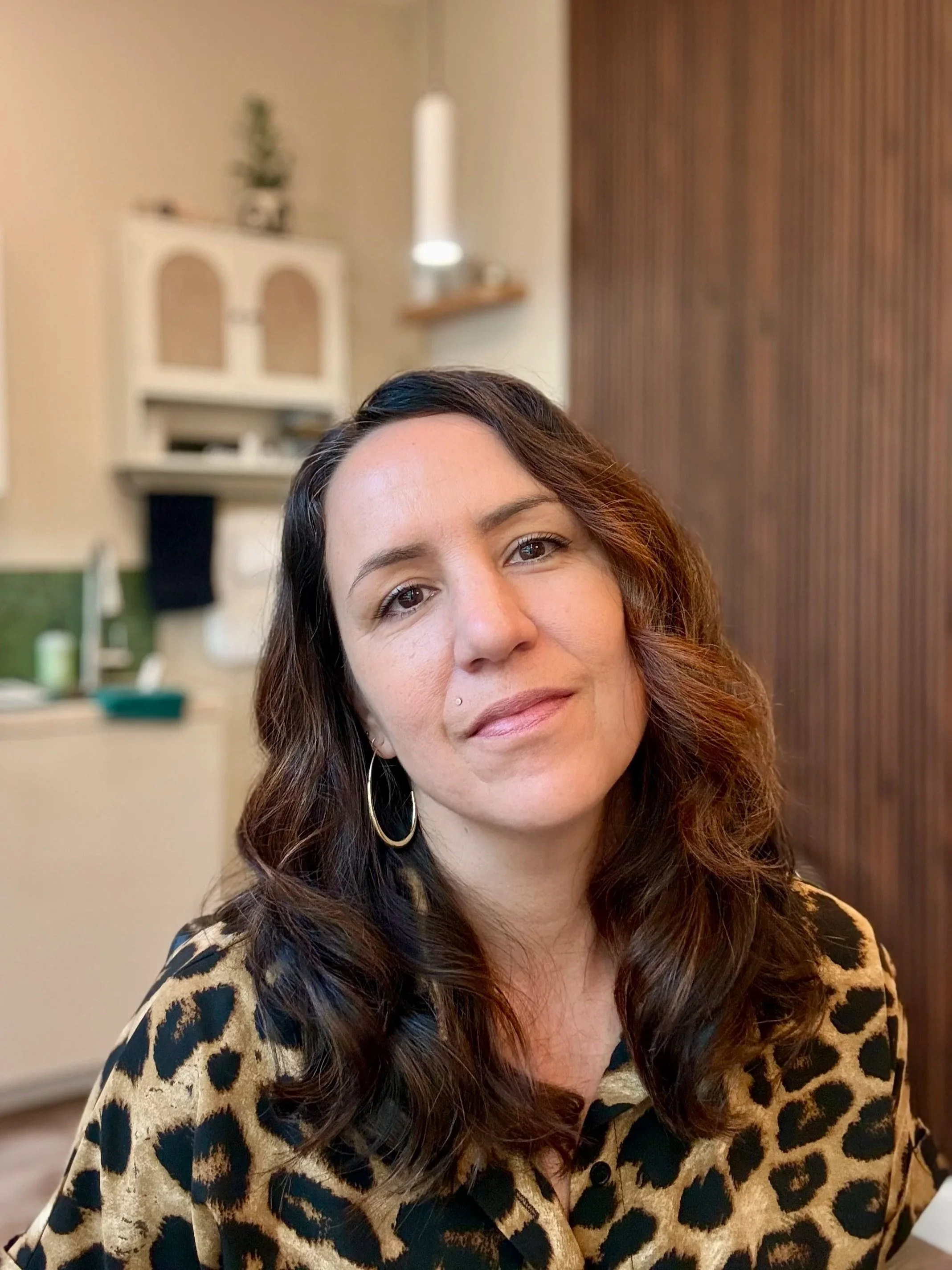 A woman with wavy brown hair and hoop earrings sitting inside a house, wearing a leopard print shirt.