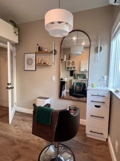 Interior view of a room with a salon chair, a white dresser, a wall mirror, a lamp, and a window with blinds, in a well-lit space.