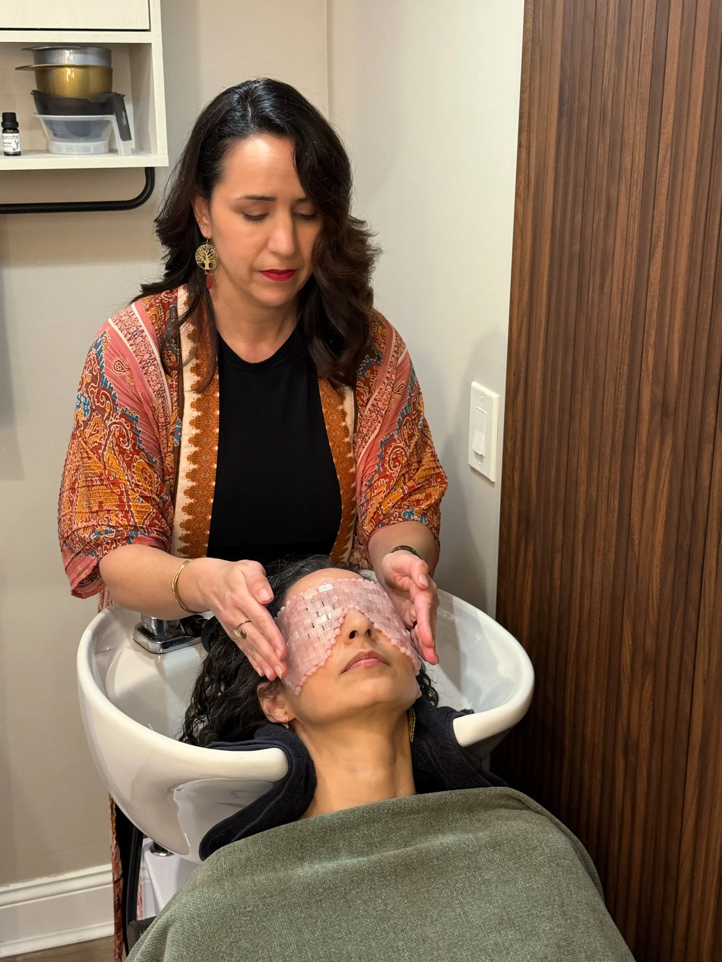 A woman receiving a facial treatment with a pink eye mask, lying in a spa bed, while a practitioner gently massages her face in a skincare clinic.