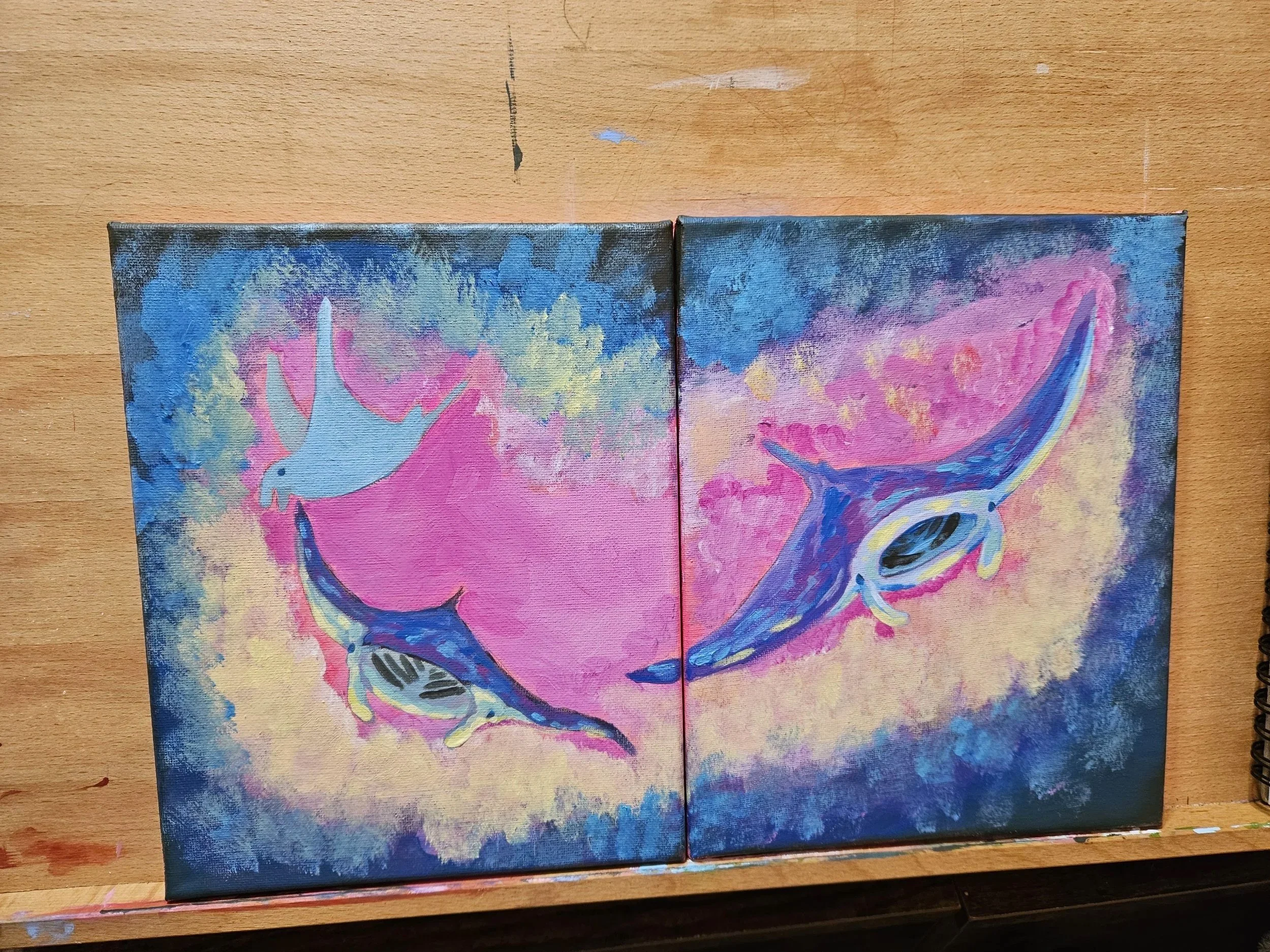 Two colorful paintings of flying whales on a wooden surface.