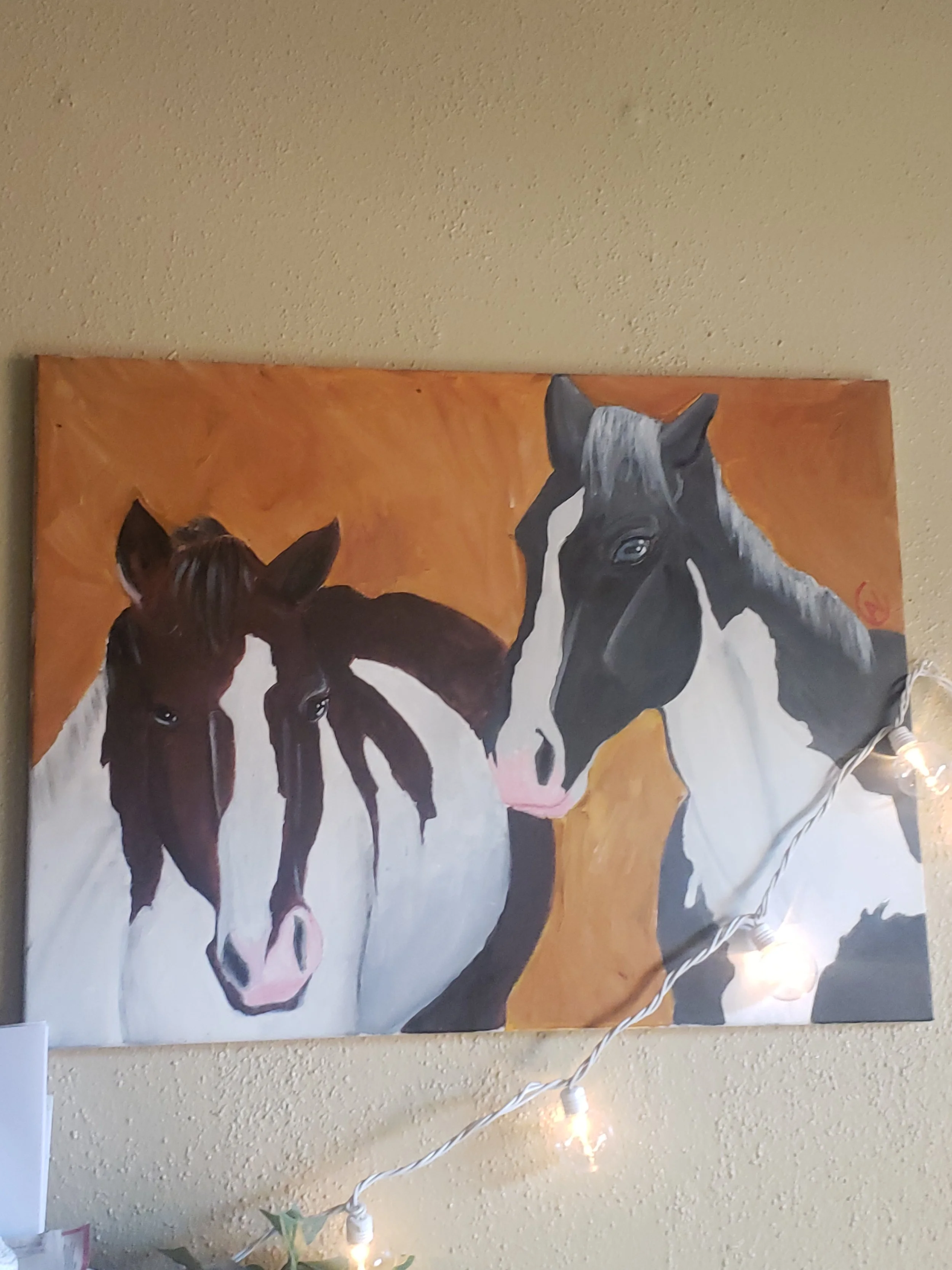 Painting of two horses, one black and white and the other black and white with brown patches, on a beige wall with string lights.