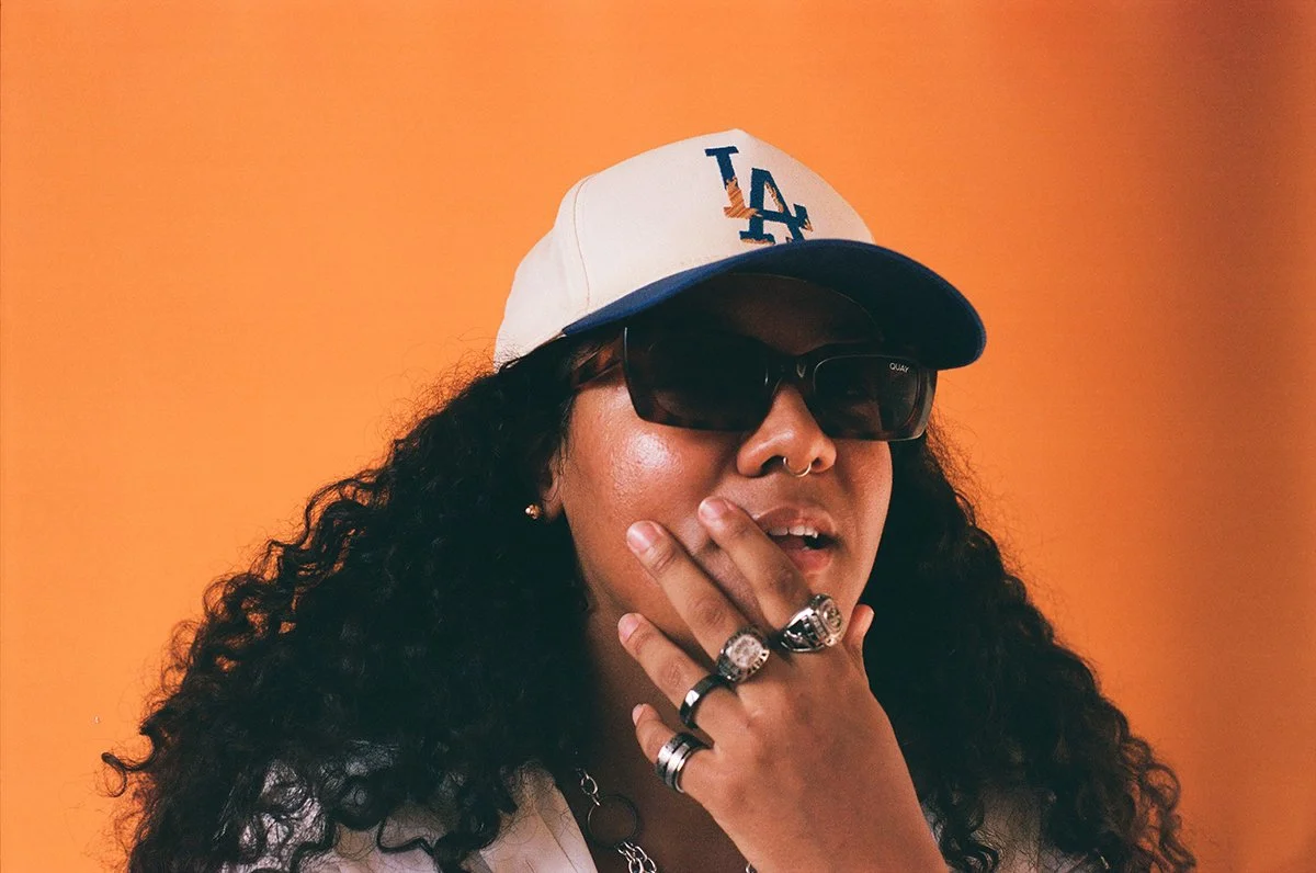 Woman with dark, long, curly hair has her hand to her face. She is wearing silver rings on all her fingers, sunglasses, and an LA baseball cap. The background is orange.