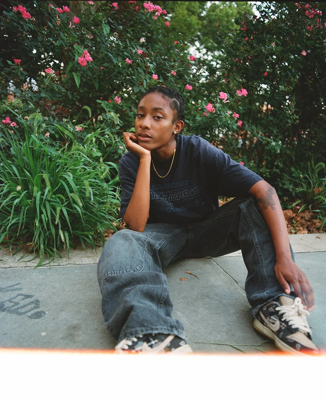 Person is sitting on the concrete ground wearing a dark blue t-shirt, grey jeans and blue nike sneakers. Their hair is braided back and they are wearing a gold chain. There are pink flowers and green plants in the background.