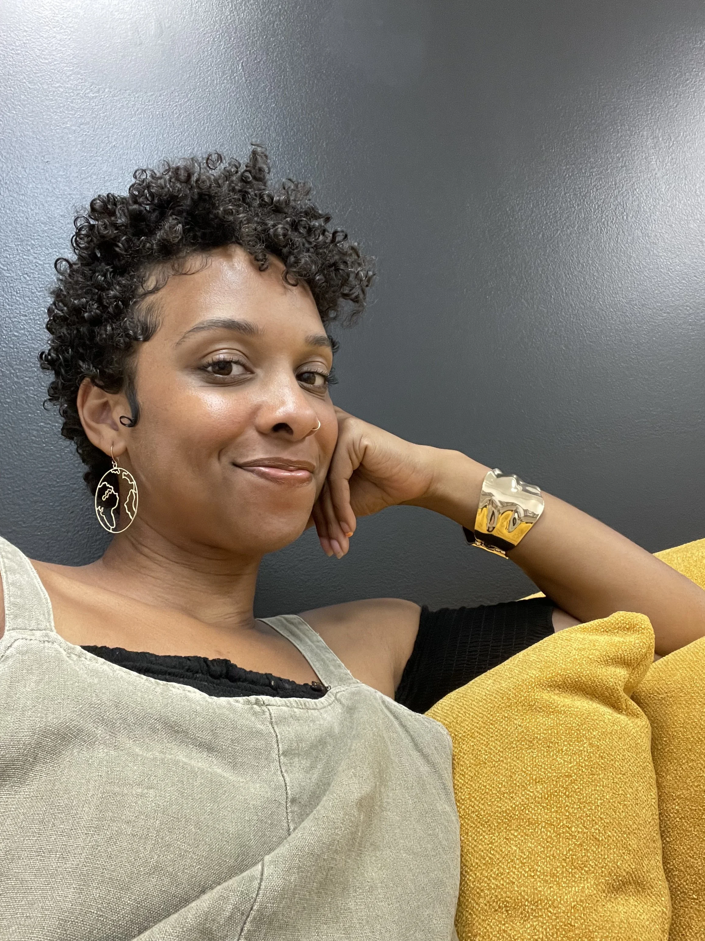 A woman with short, curly hair and wearing earrings with a face outline, a large gold cuff bracelet, a black top with cut-out shoulders, a beige sleeveless dress, sitting on a yellow cushion against a dark gray wall.