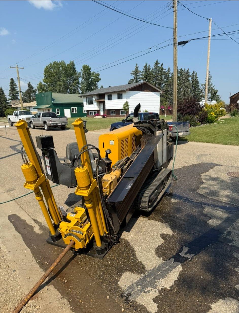 A small tracked drilling machine positioned on a street, with buildings, parked trucks, and power lines in the background on a clear day.