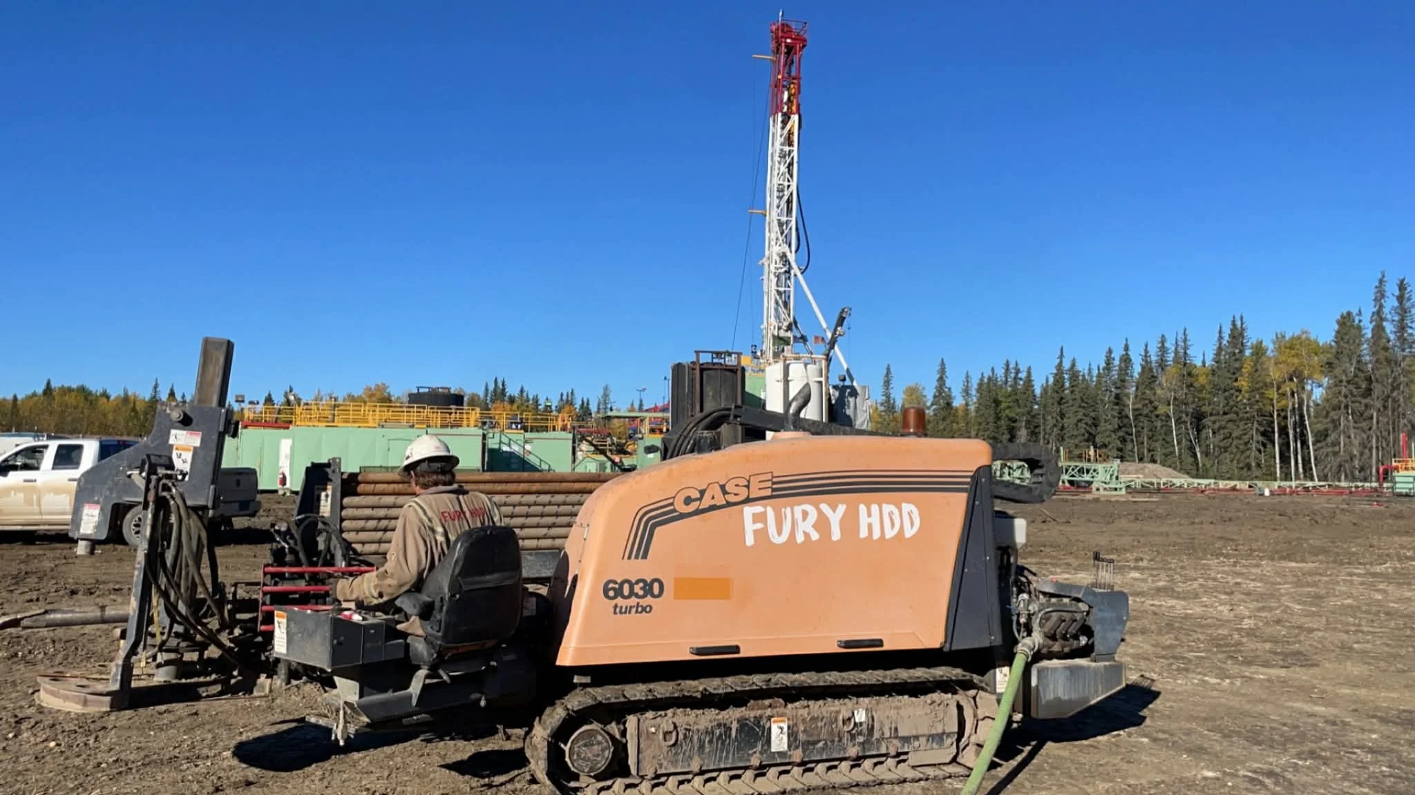 Construction worker operating a CASE FURY HDD 6030 turbo horizontal boring machine on a construction site with dirt ground, pipes in the background, and a drilling tower against a clear blue sky.