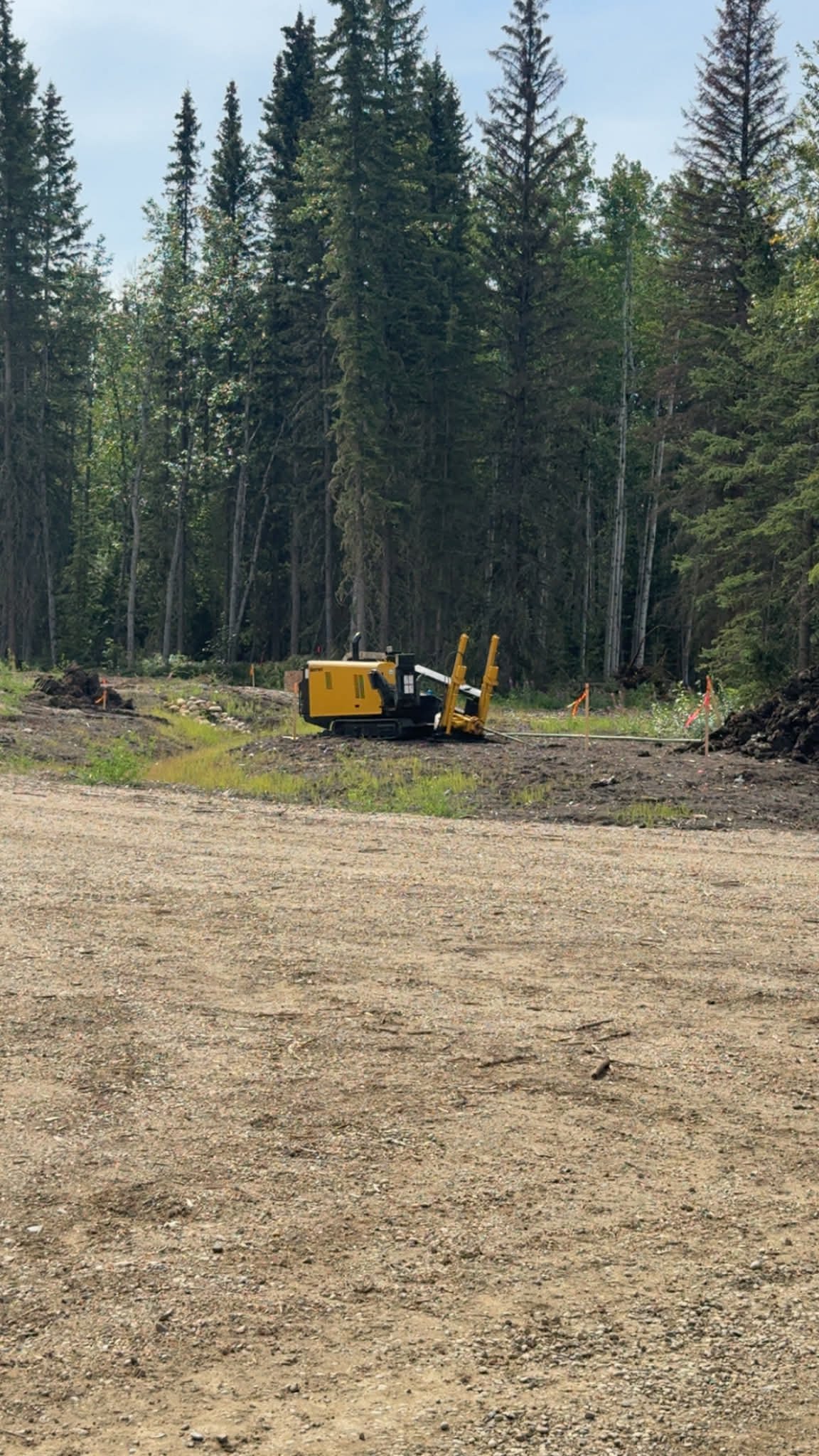 A construction site in a forest with a yellow piece of heavy machinery, possibly a trencher, on a dirt path surrounded by tall trees and orange safety flags.