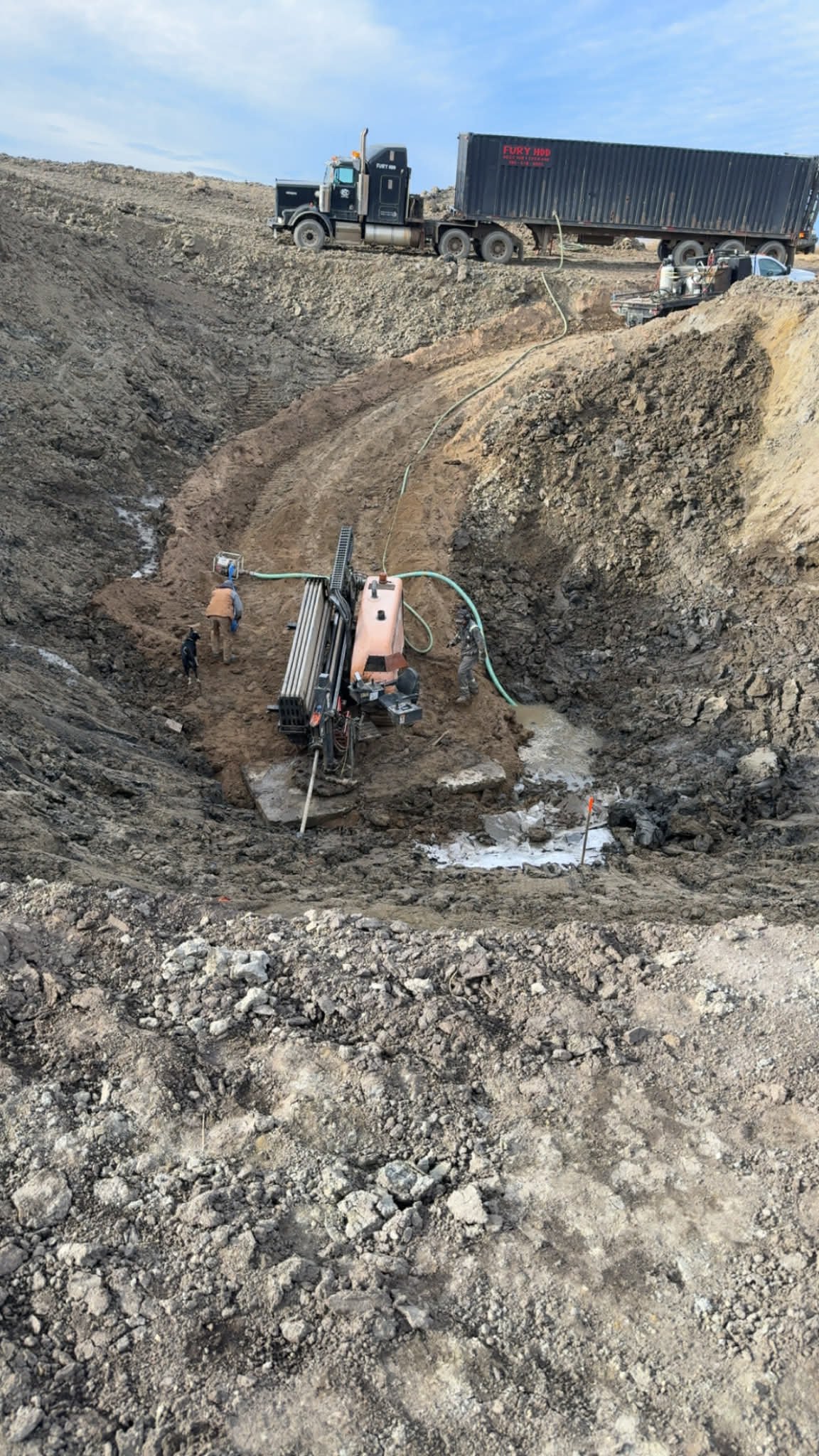 Construction site with workers operating equipment in a large excavated trench, truck parked on the edge of the excavation.