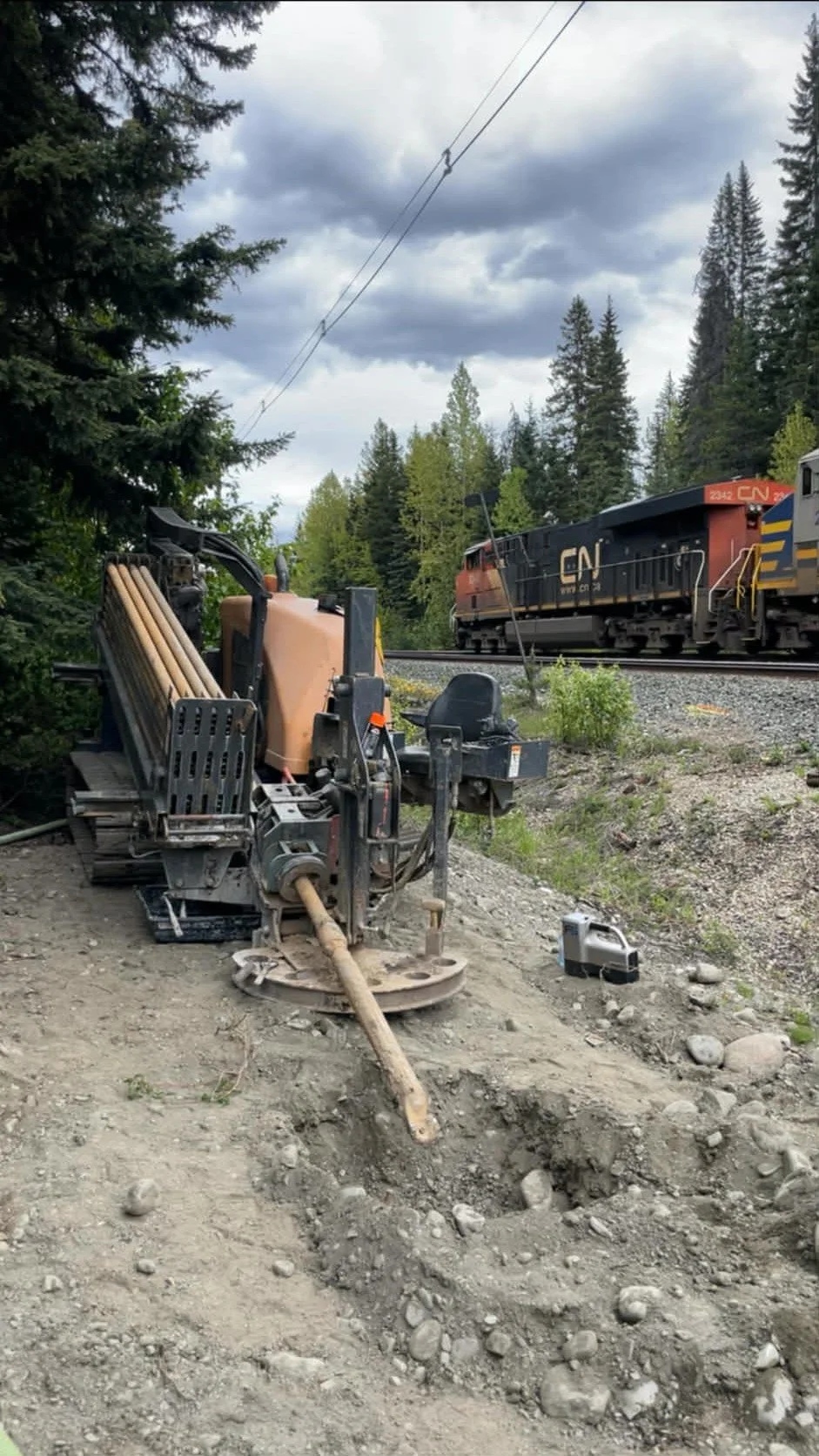 Construction equipment on dirt beside a railroad track with a train passing by in a forested area under cloudy sky.