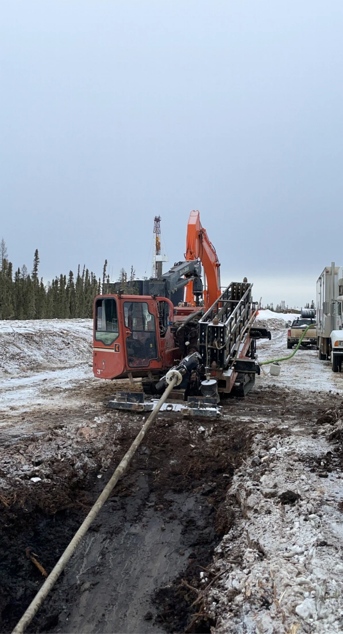 A construction site with an excavator digging a trench in snowy terrain, with trees and construction vehicles in the background.