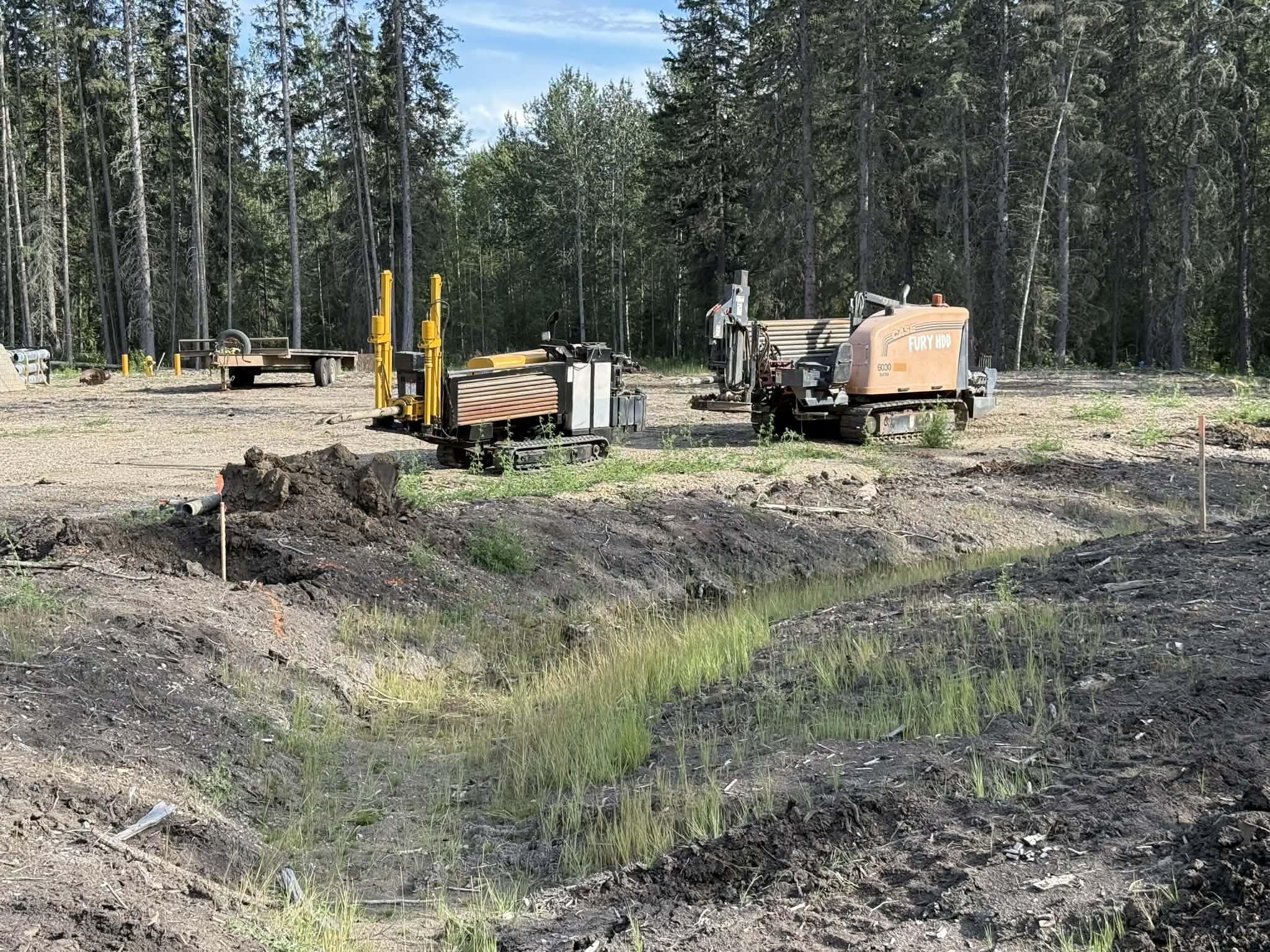 Construction site with two small bulldozers, one labeled 'Fury HDD,' on disturbed land with a small waterway in the foreground. Trees line the background.