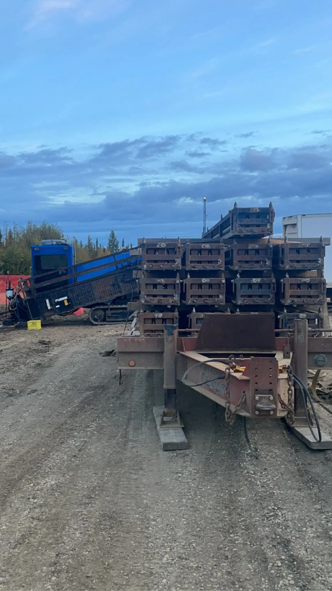 Construction site with a trailer carrying metal beams and a blue equipment vehicle in the background under a cloudy sky.