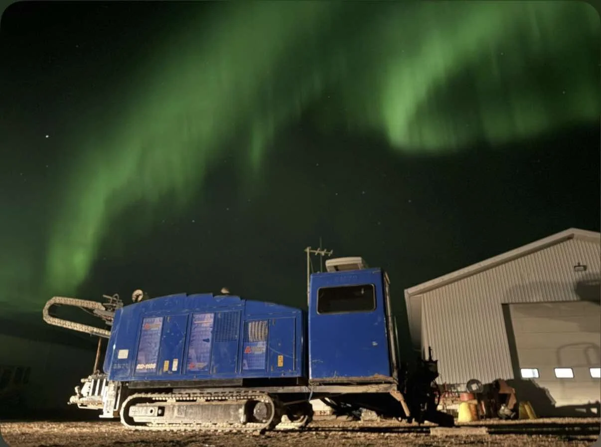 A large blue horizontal directional drill machine with tracks, parked outside a white building at night, under the green Northern Lights in the sky.