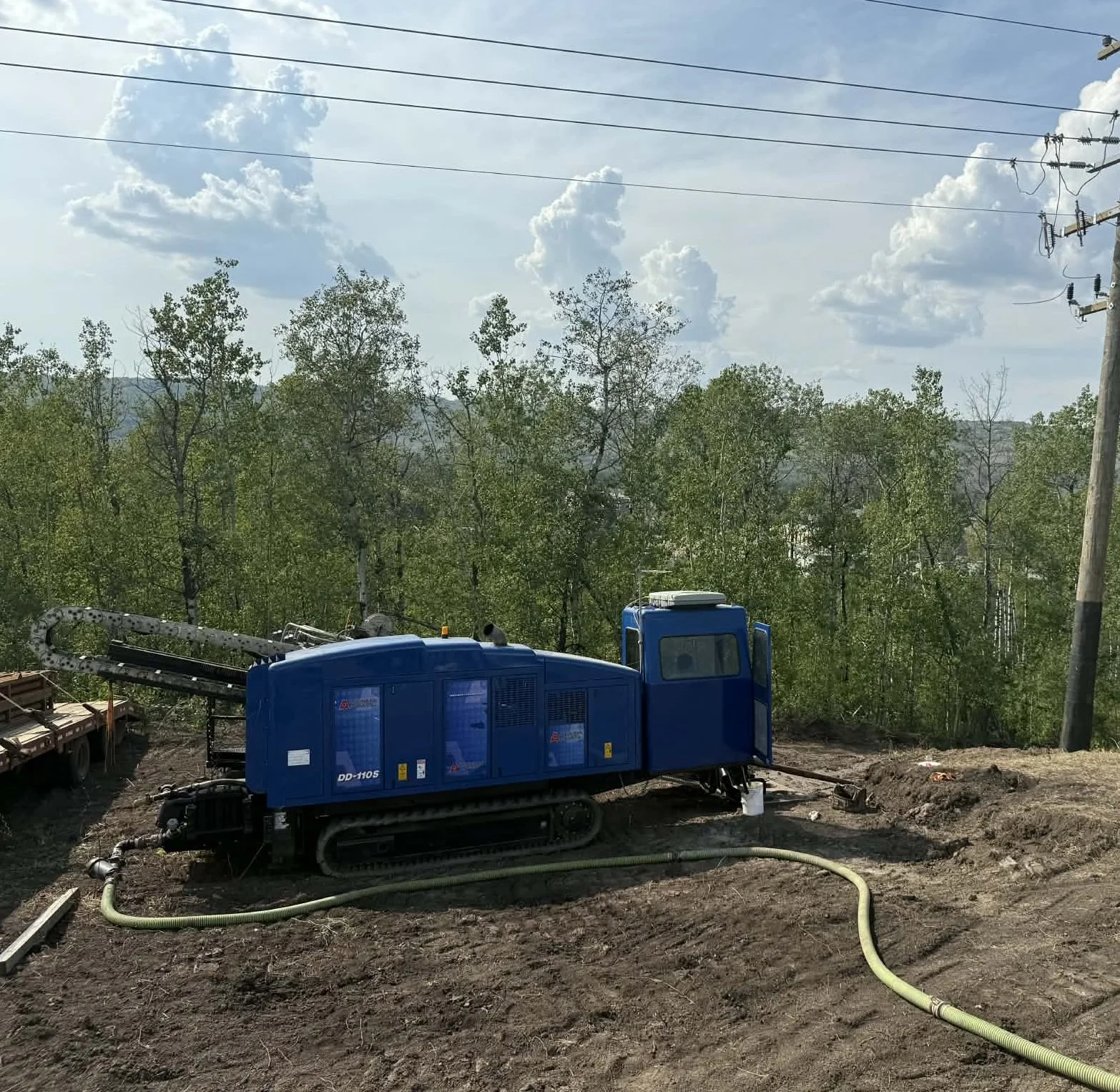 A blue industrial machine on tracks operating on a dirt site, connected to a yellow flexible hose with infrastructure and trees in the background, under a partly cloudy sky.