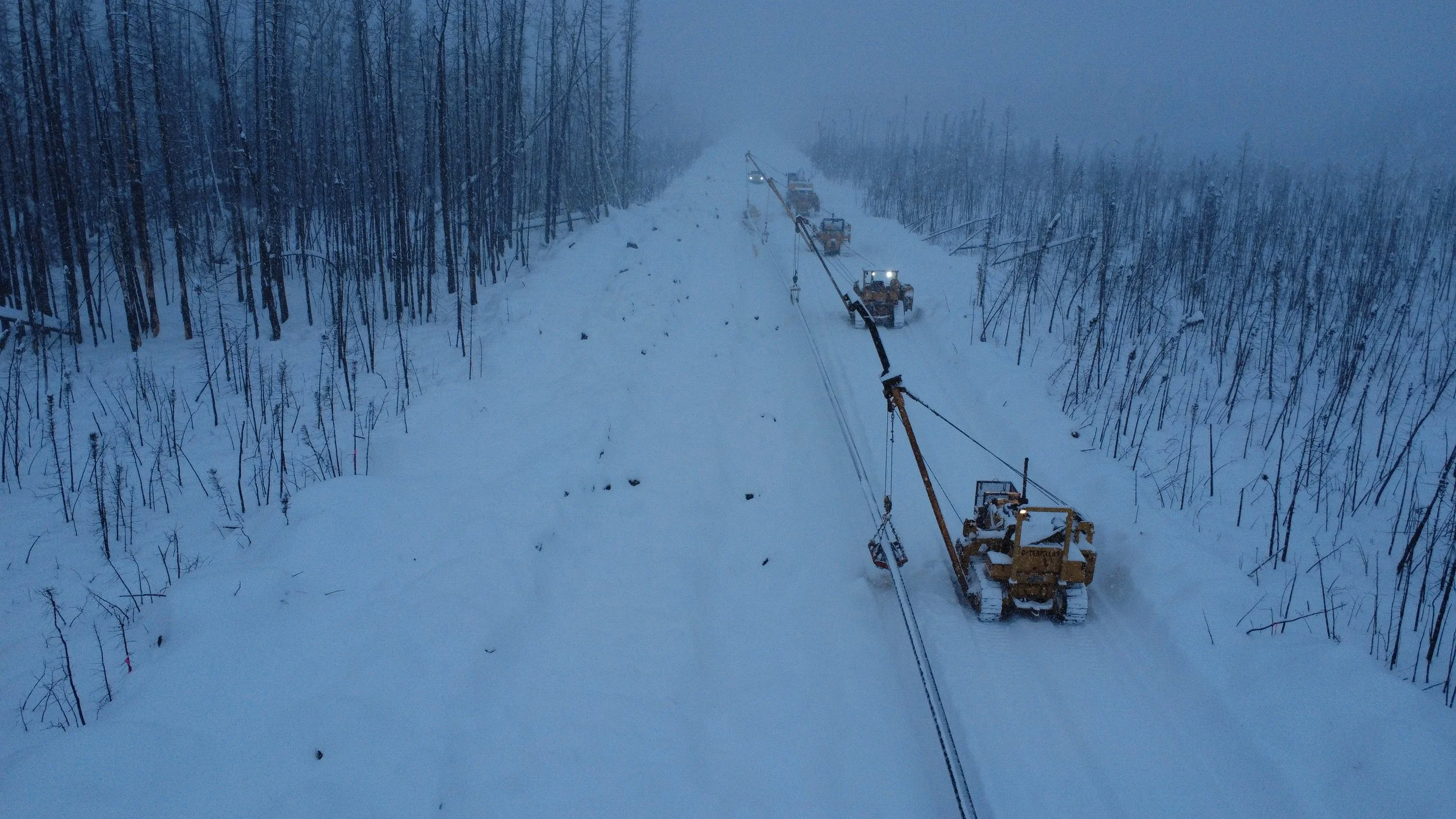 Snow-covered road being cleared by multiple large bulldozers and equipment, flanked by leafless trees, with foggy and cold atmosphere.