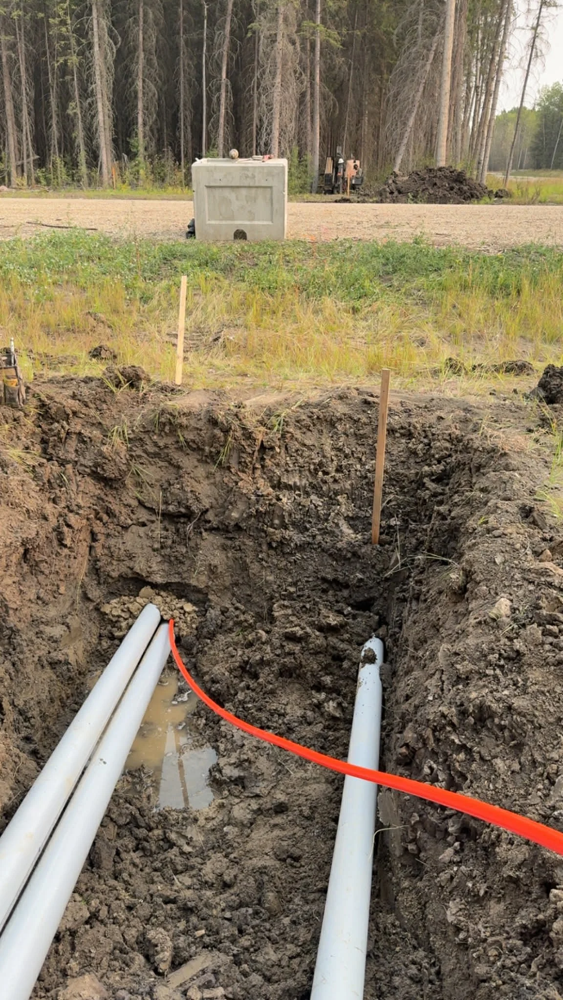 Underground utility pipes being installed in a trench at a construction site, with a wooded background and construction equipment in the distance.