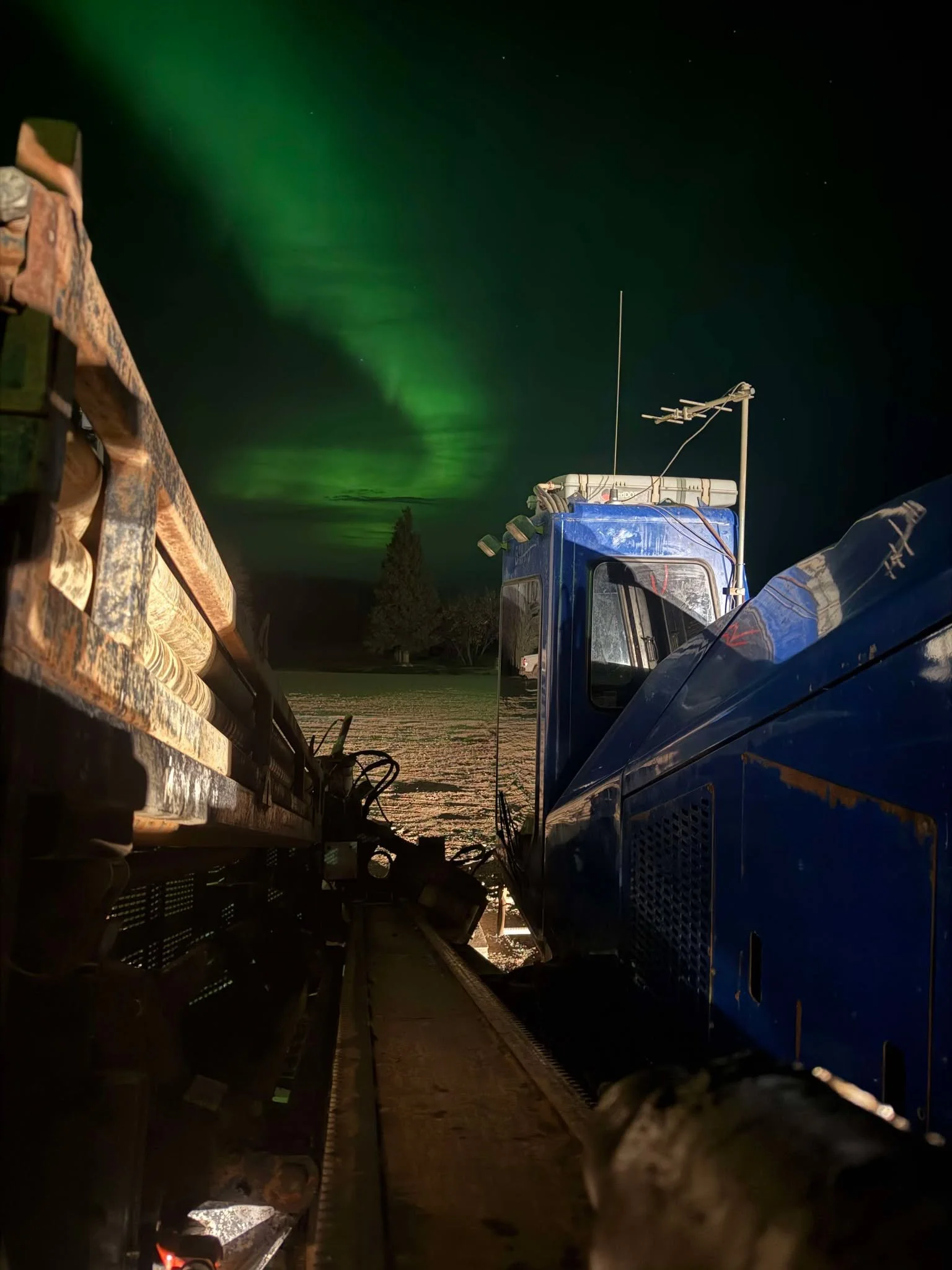 Photo taken from the ground level of a blue tractor or heavy machinery at night, with the northern lights (aurora borealis) visible in the sky, illuminating greenish clouds, with a silhouette of a tree in the distance.