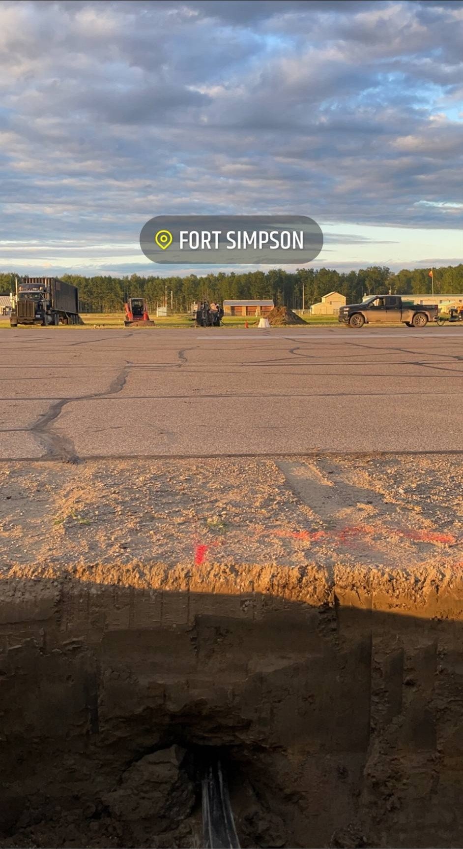 View of a construction site at Fort Simpson with pipes being installed underground, machinery, trucks, and workers present, under a cloudy sky.