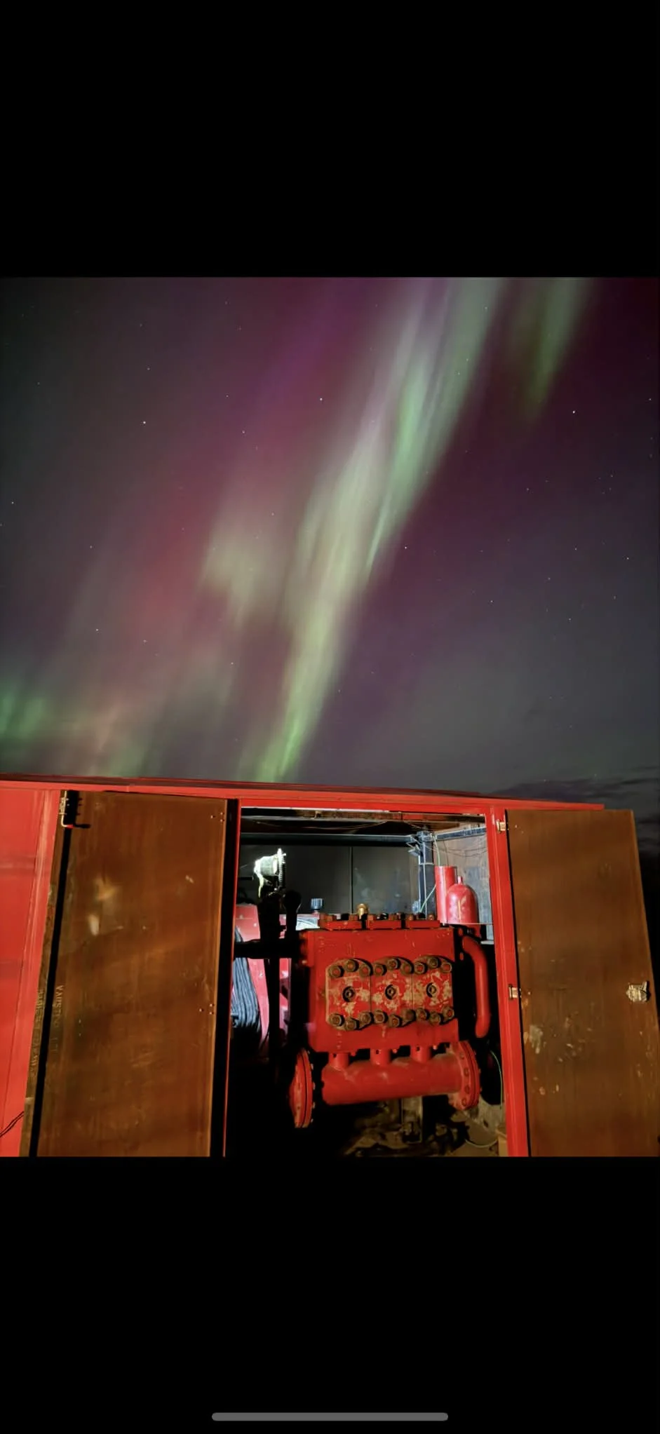 Night sky with green and purple aurora borealis above a red building with machinery inside.