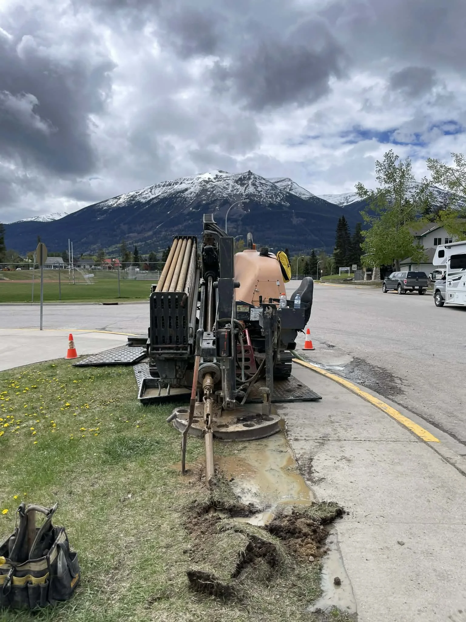 Road construction equipment working on a sidewalk with snow-capped mountains in the background, cloudy sky overhead, traffic cones nearby, and parked cars.