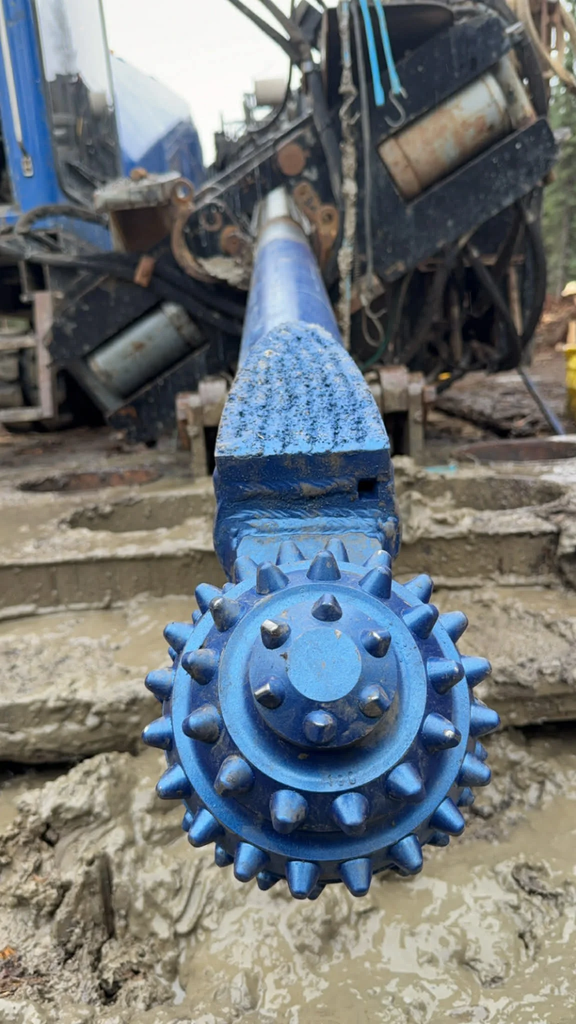 Close-up of a large blue drill bit attached to a piece of heavy construction equipment, with a muddy construction site background.